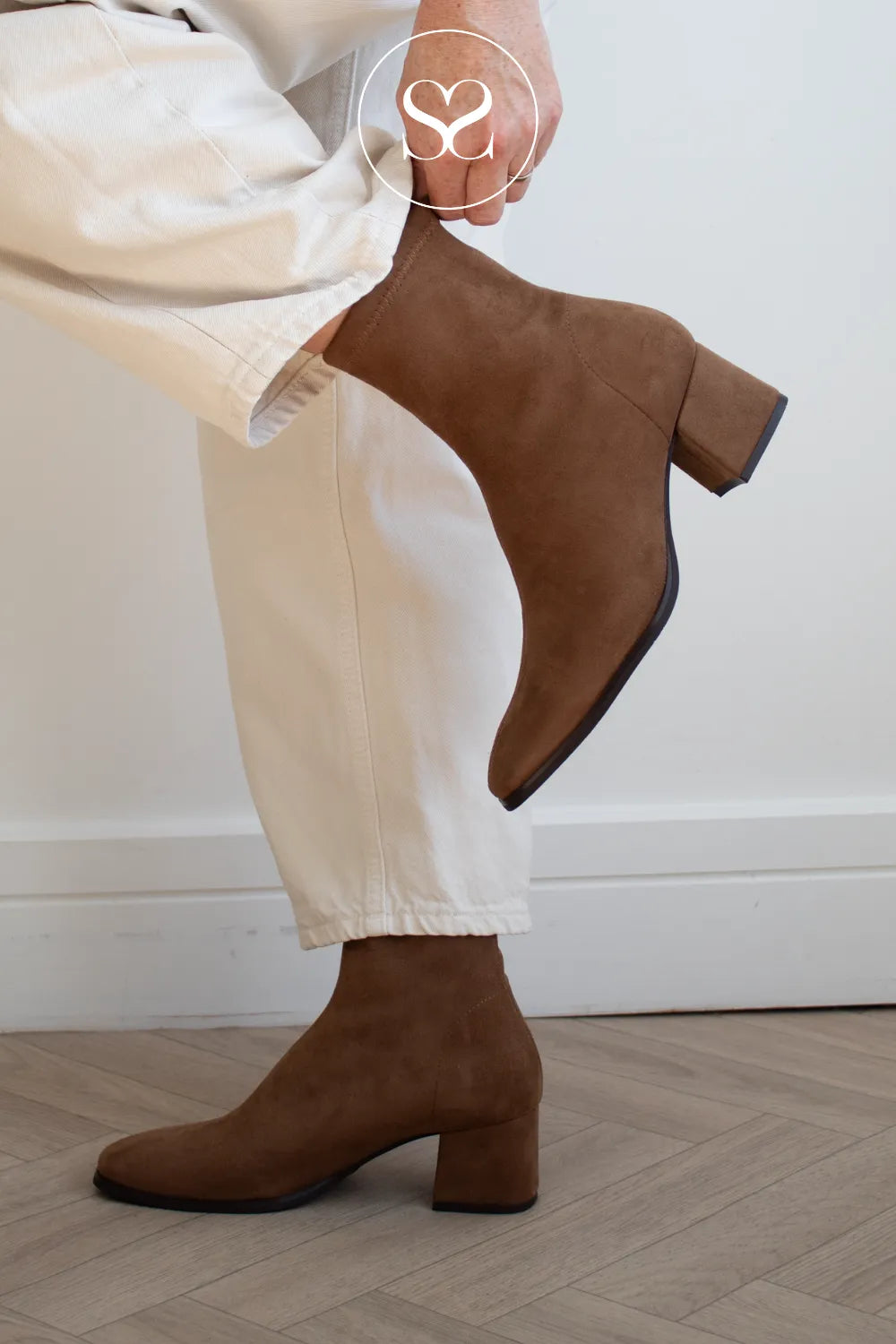 A person wearing Miss Elastic brown suede ankle boots with block heels and white trousers, shown from the waist down against a light wall and wood floor.