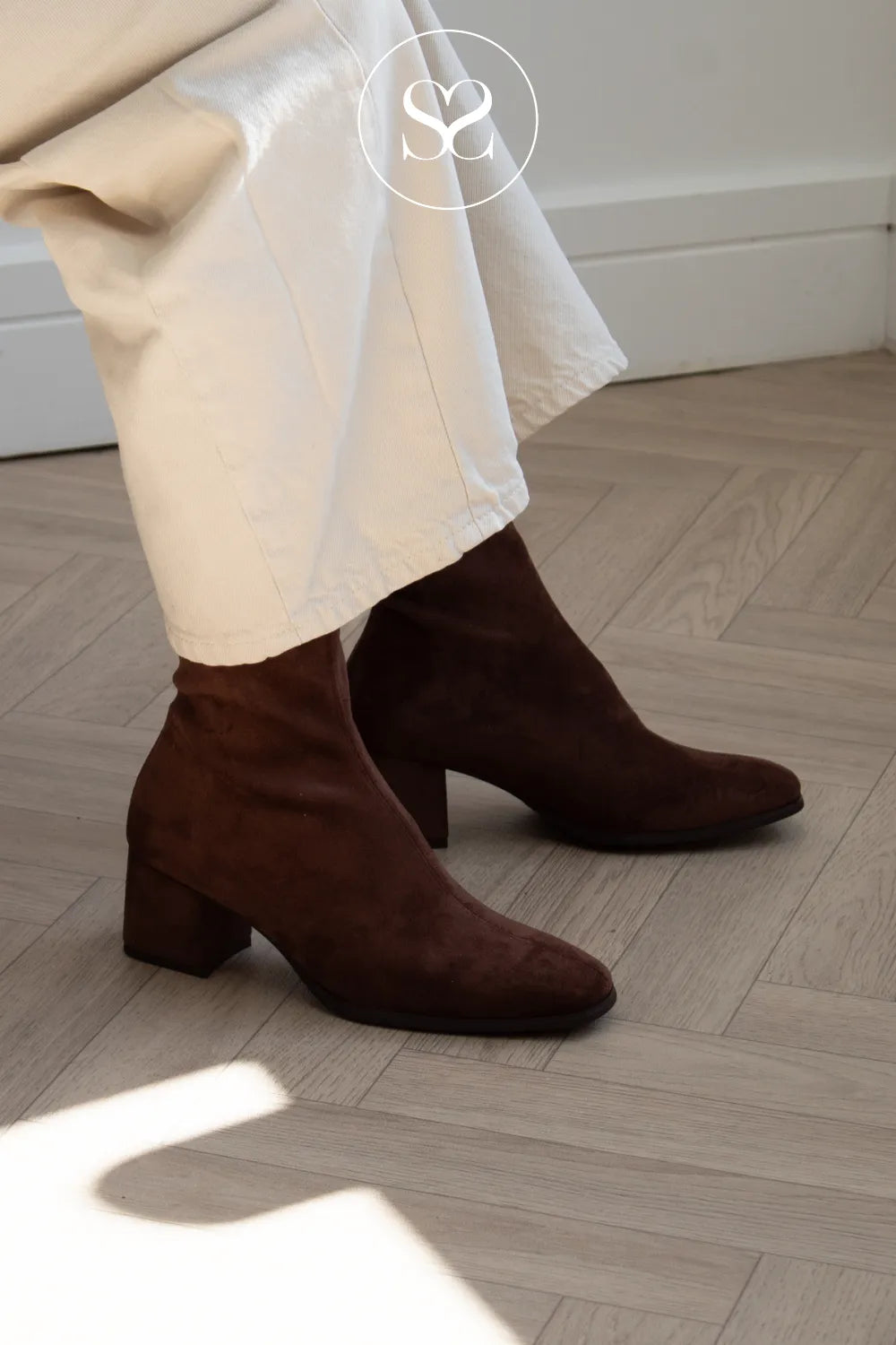 A pair of Miss Elastic dark brown suede ankle boots with block heels, worn with white trousers on a light wood herringbone floor in Ireland