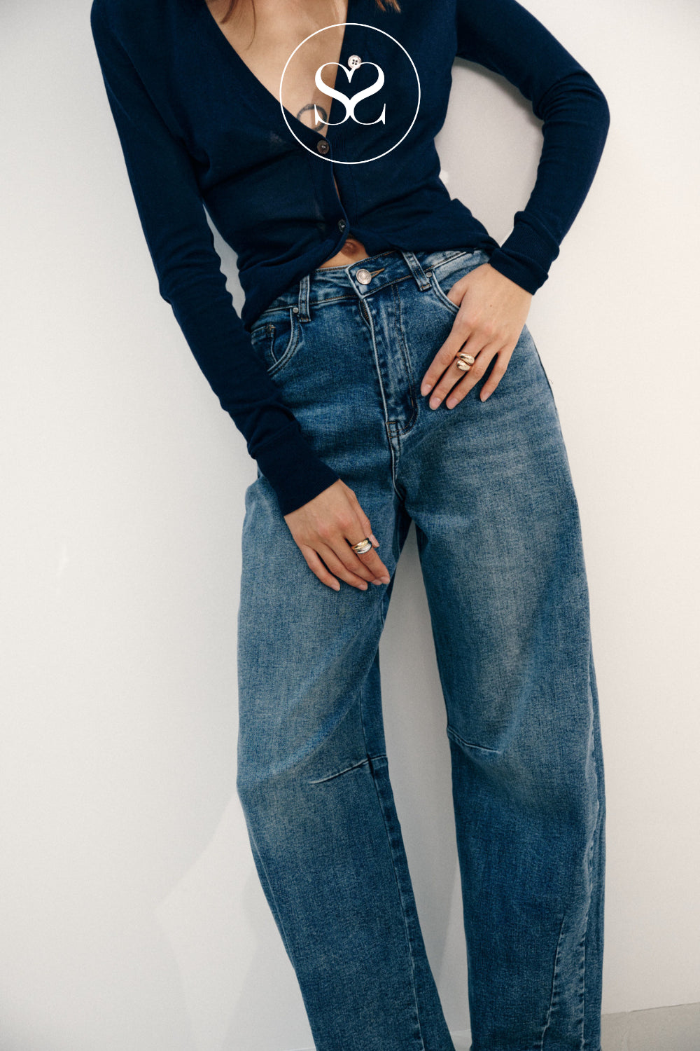 A model wearing a navy blue long-sleeve knit top and high-waisted, wide-leg blue denim jeans, standing against a plain off-white wall.