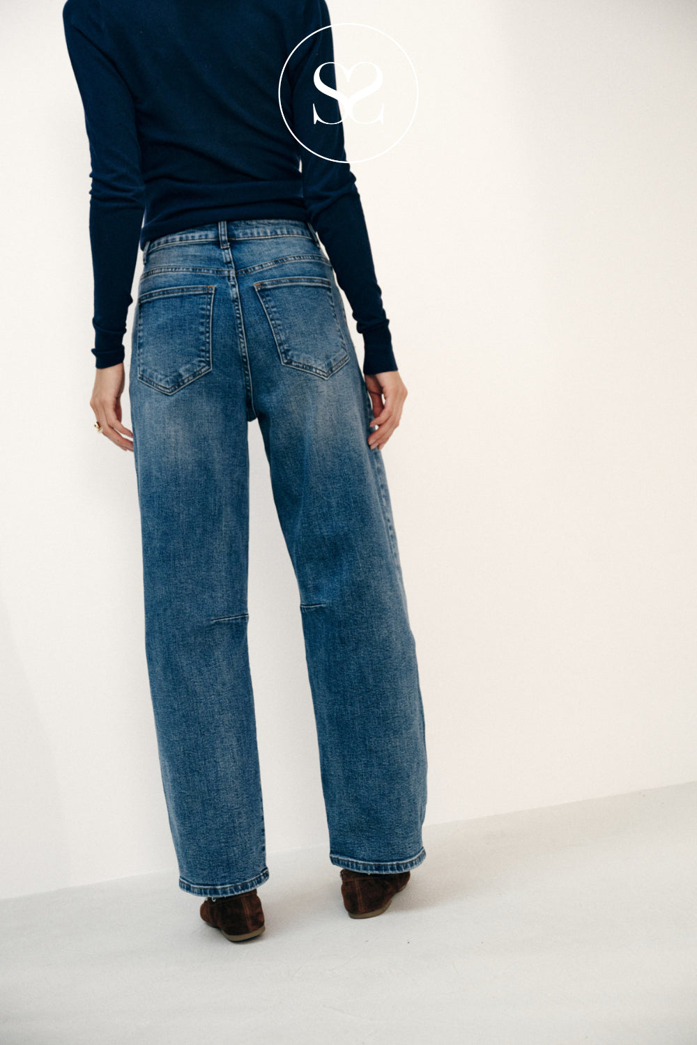 A model shown from the back wearing high-waisted, wide-leg blue denim jeans with a dark navy long-sleeve top and brown shoes, set against a plain white studio background.