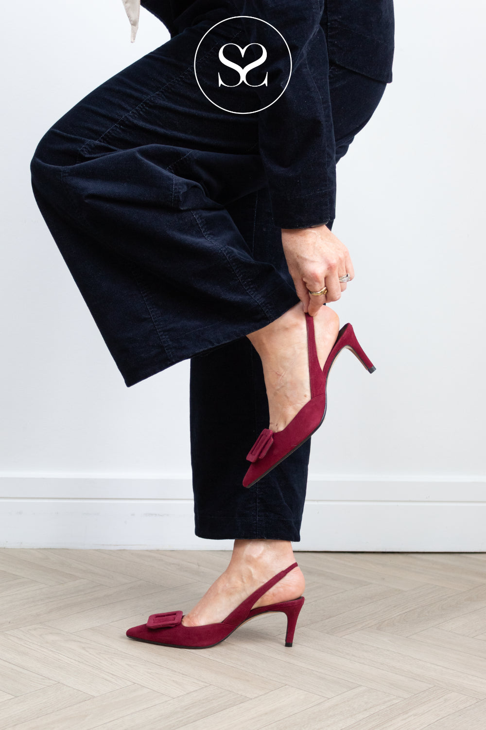 A woman wearing dark navy velvet trousers and burgundy slingback kitten heeled shoes with buckle details, shown from the waist down in a room with a light gray wall and wood floor. Shoes are Lodi nestor in Burgundy Red suede