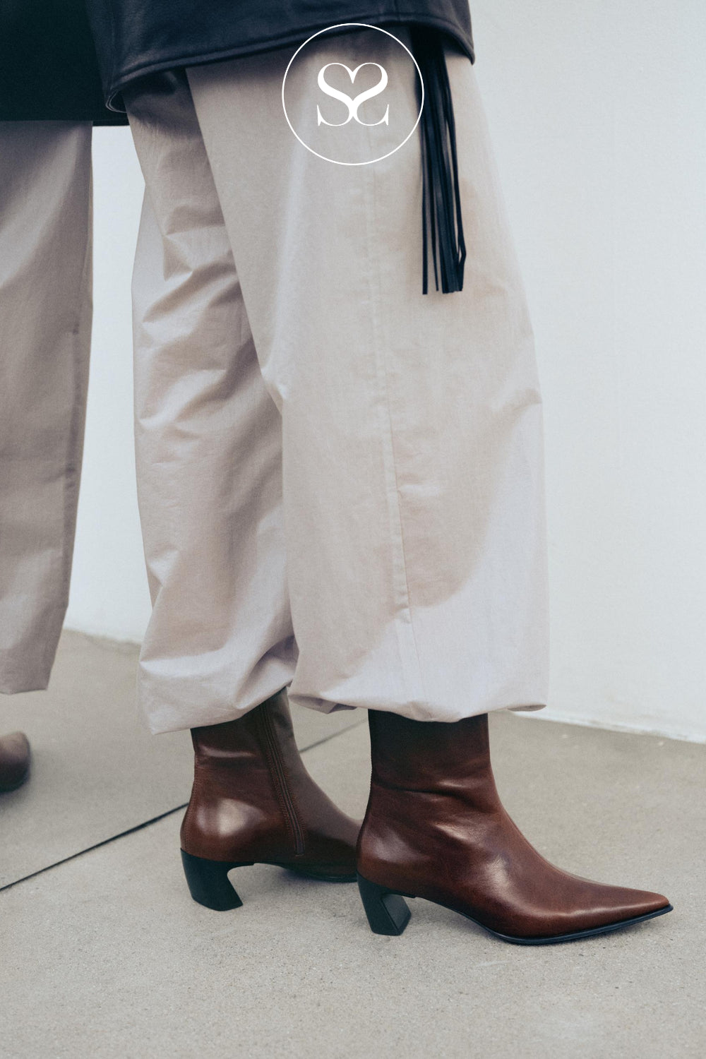 A close-up lifestyle shot of a person wearing brown leather pointed-toe ankle boots with low curved block heels from Vagabond shoemakers paired with light beige wide-leg trousers and a black tassel belt, standing against a plain light gray wall.