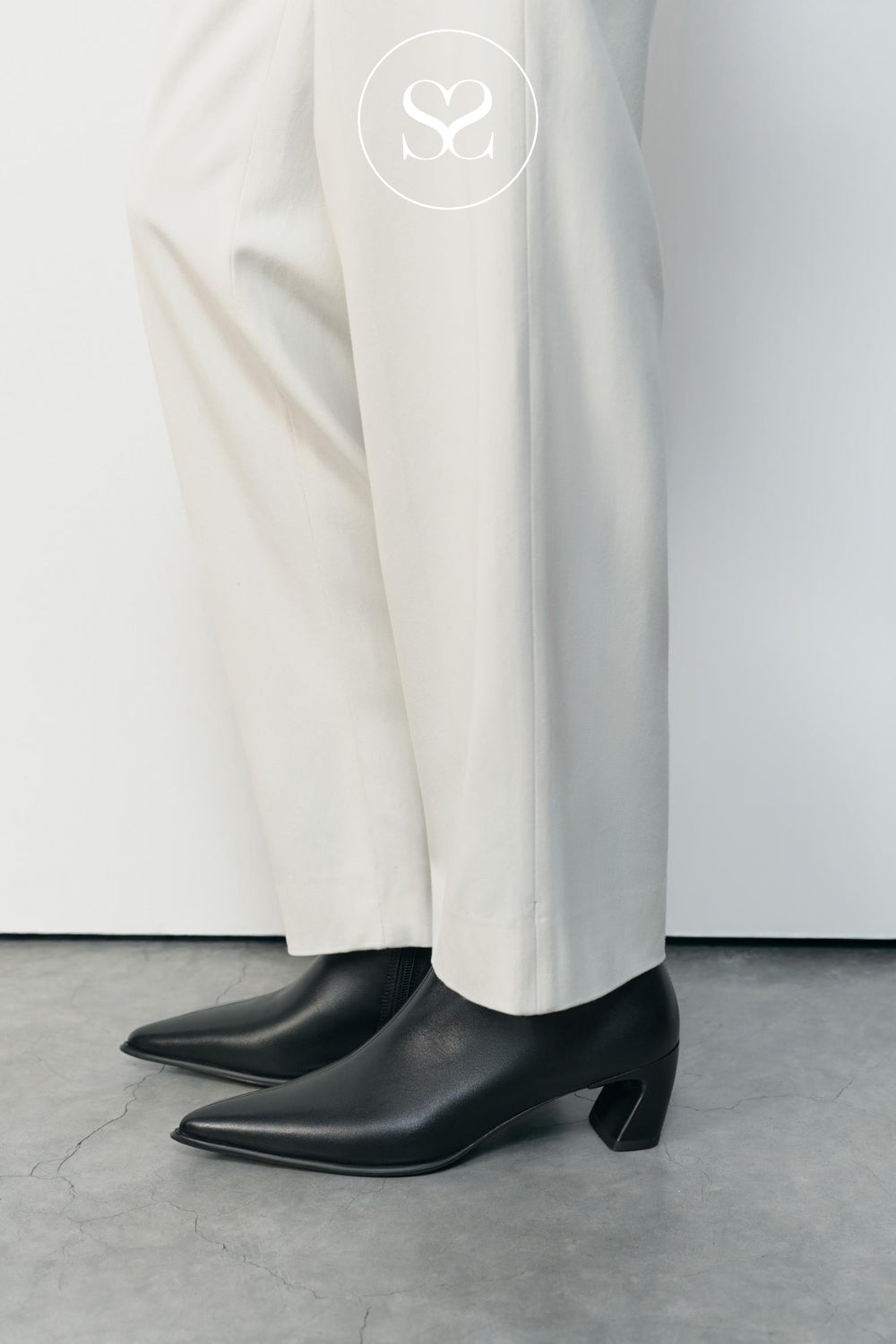 Sleek black pointed-toe ankle boots with a curved block heel from Vagabond, shown on a model wearing off-white trousers against a minimalist studio background.