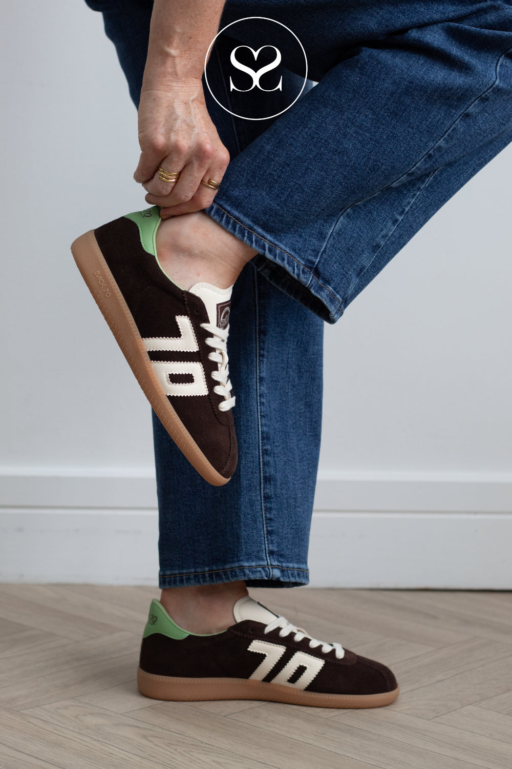 An Irish woman wearing blue jeans and brown suede sneakers from Back 70' lettering, white laces, and a light green heel tab, standing on a light wood floor against a white wall.