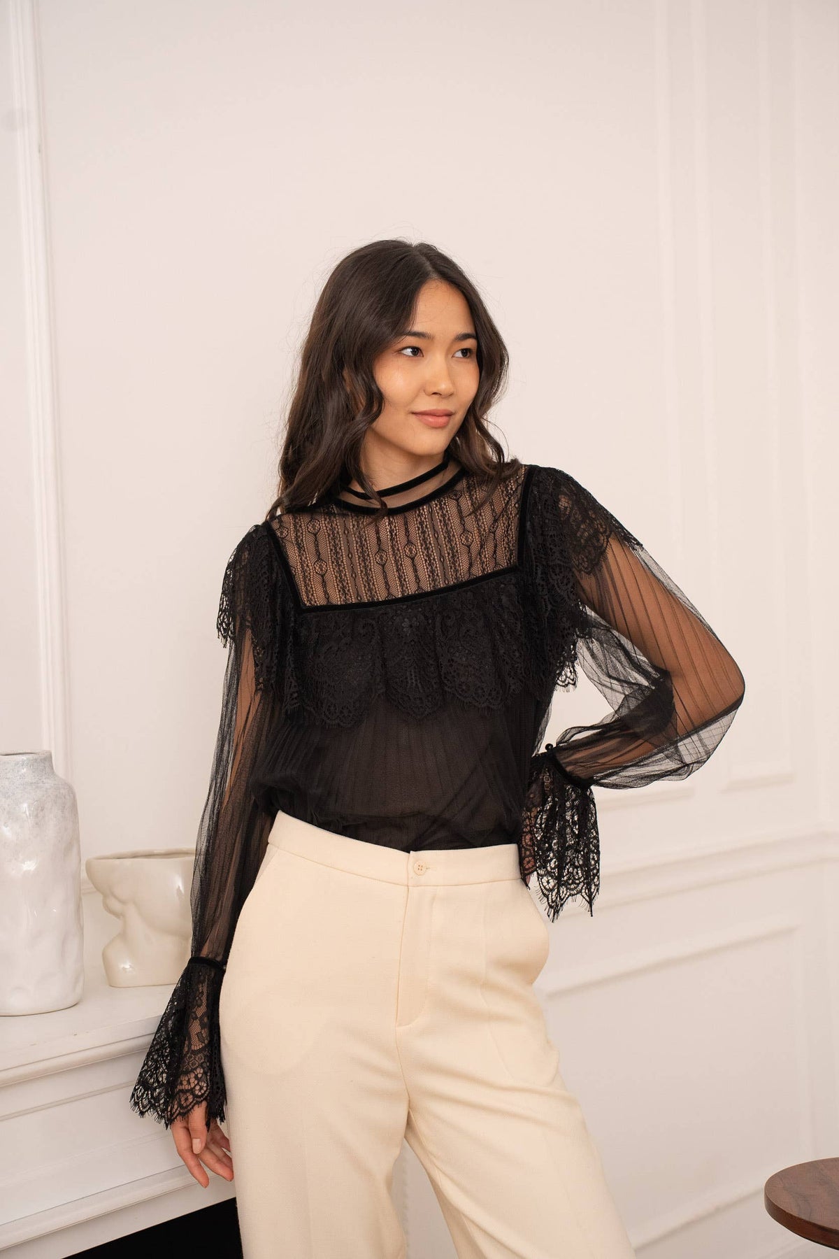 Woman wearing a black sheer blouse with a textured top and white pants indoors.