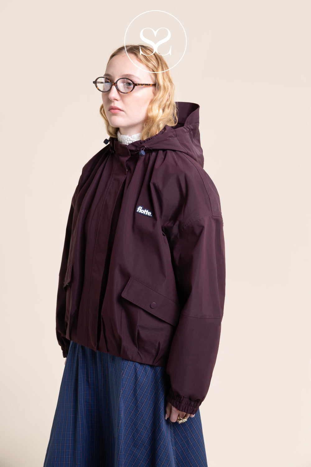 A model wearing a dark burgundy Flotte hooded jacket with a small white logo on the chest, paired with a blue pleated skirt, set against a solid beige background.