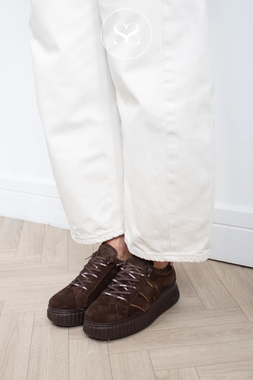 A person wearing Wonders comfortable dark brown suede platform sneakers with white pants, standing on a light wood herringbone floor against a white wall.