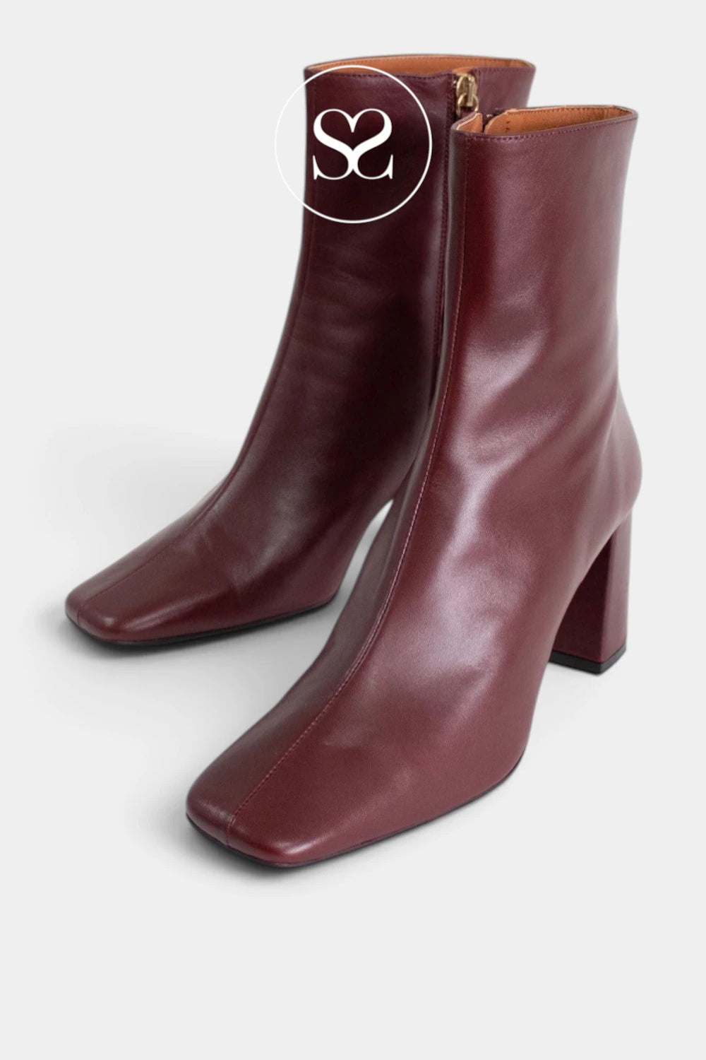 A pair of burgundy leather ankle boots with a block heel and square toe, shown on a light gray background. A white circular logo overlay with 'SS' is visible on the upper boot.