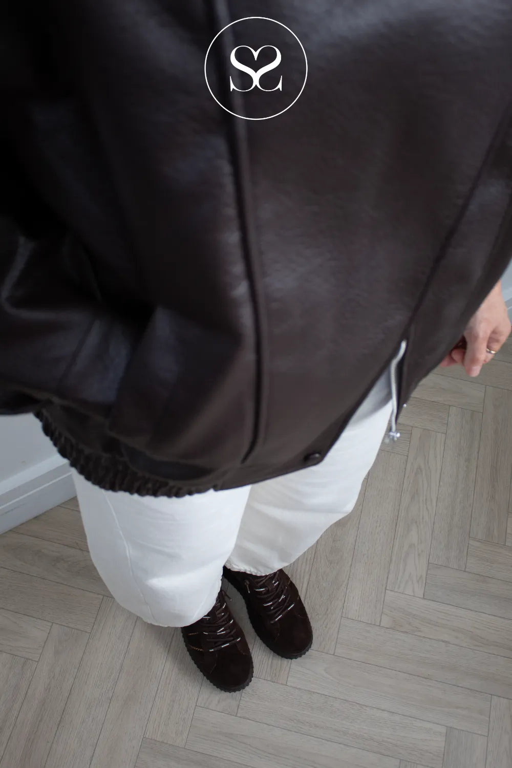 A person wearing a dark brown leather jacket, white pants, and dark brown lace-up platfrom trainers standing on a light wood herringbone floor. A circular logo overlay is visible at the top center.