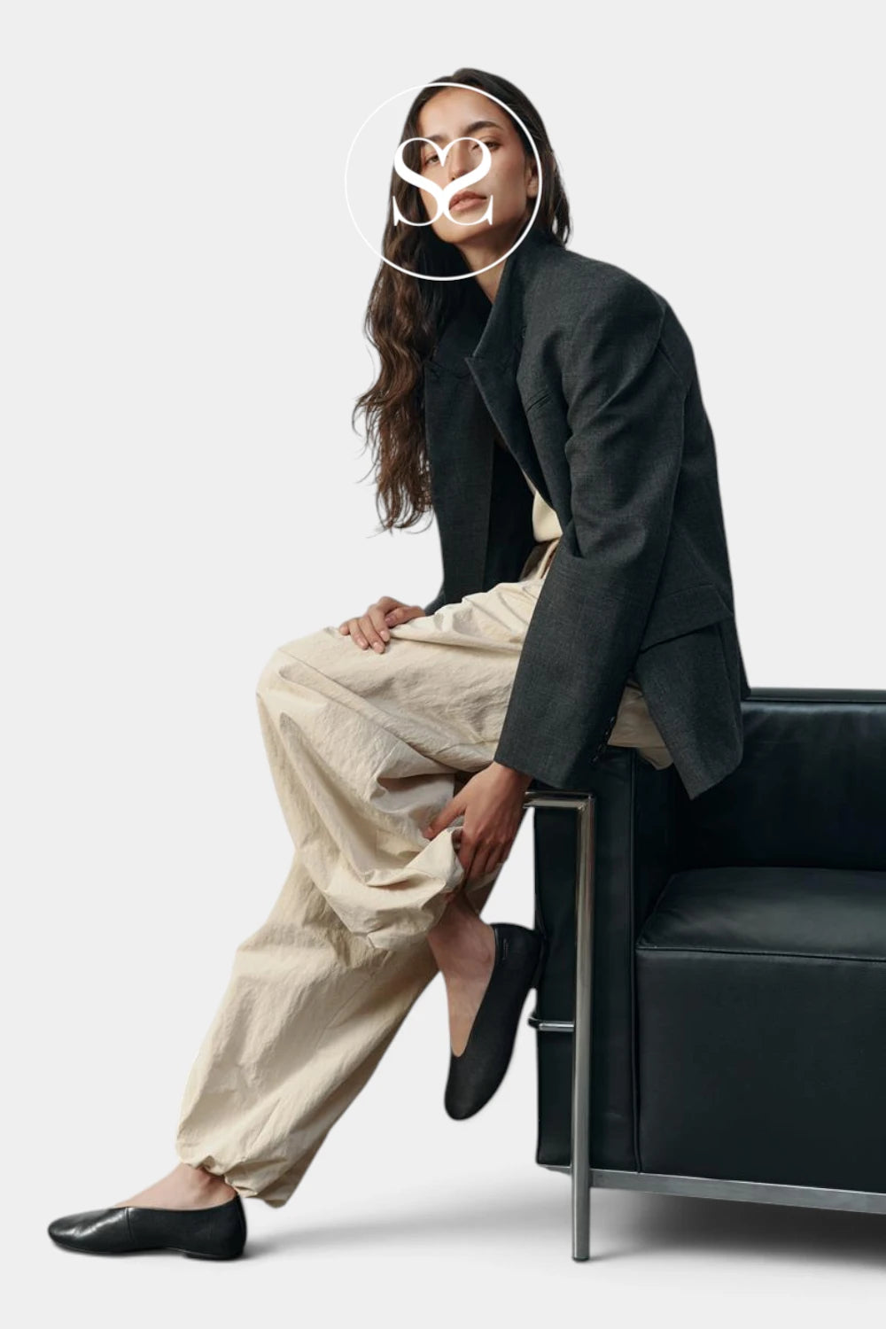 A woman sits on a black leather chair wearing a dark charcoal blazer, beige wide-leg trousers, and black flats against a light gray studio background.