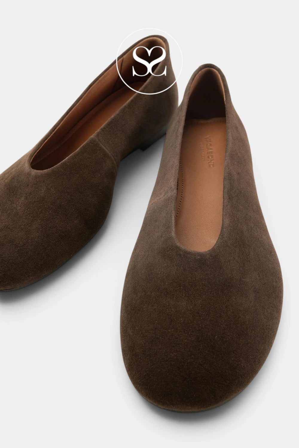 A pair of dark brown suede slip-on shoes with a low block heel, displayed on a light gray background. One shoe is positioned upright to show the interior, while the other lies flat at an angle.