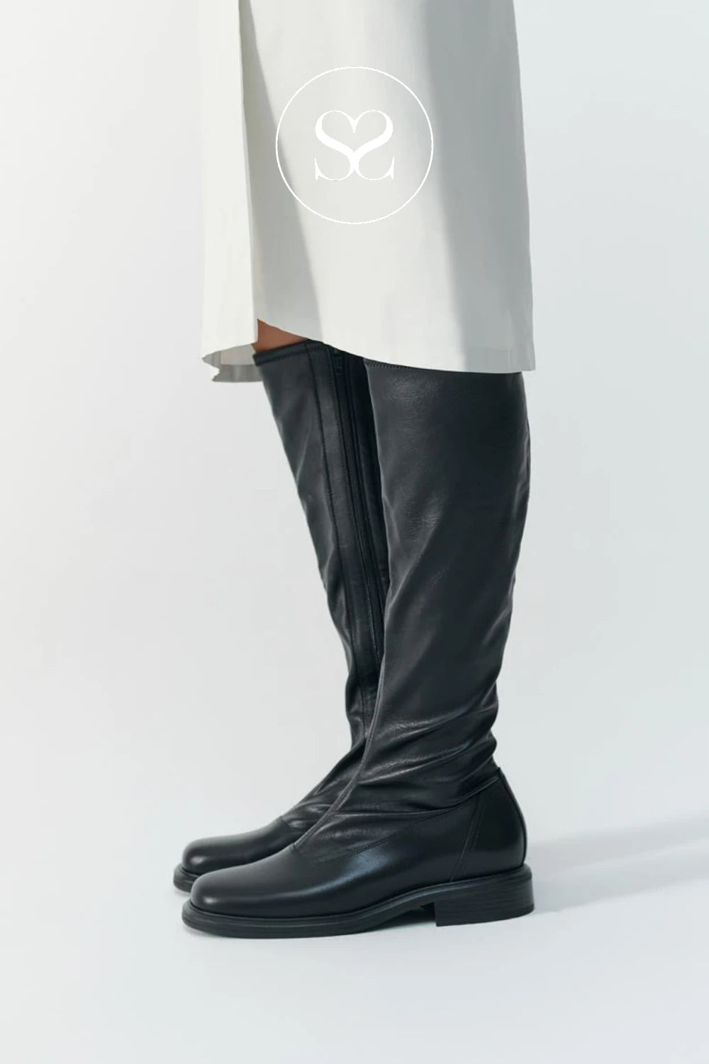 A pair of black leather knee-high boots with a side zipper, shown on a model wearing a white skirt. A circular logo with an 'S' inside a circle is visible on the white fabric.
