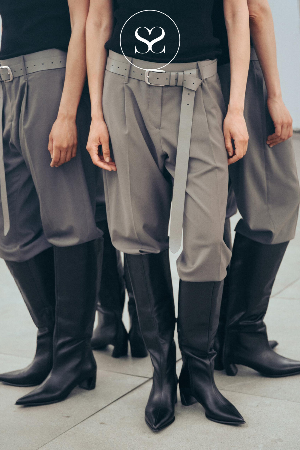 Three models standing side-by-side wearing grey wide-leg cropped trousers with matching grey belts and Vagabond black pointed-toe knee-high boots. The center model has a black top with a circular 'Sheneil Shoes' logo on the chest.
