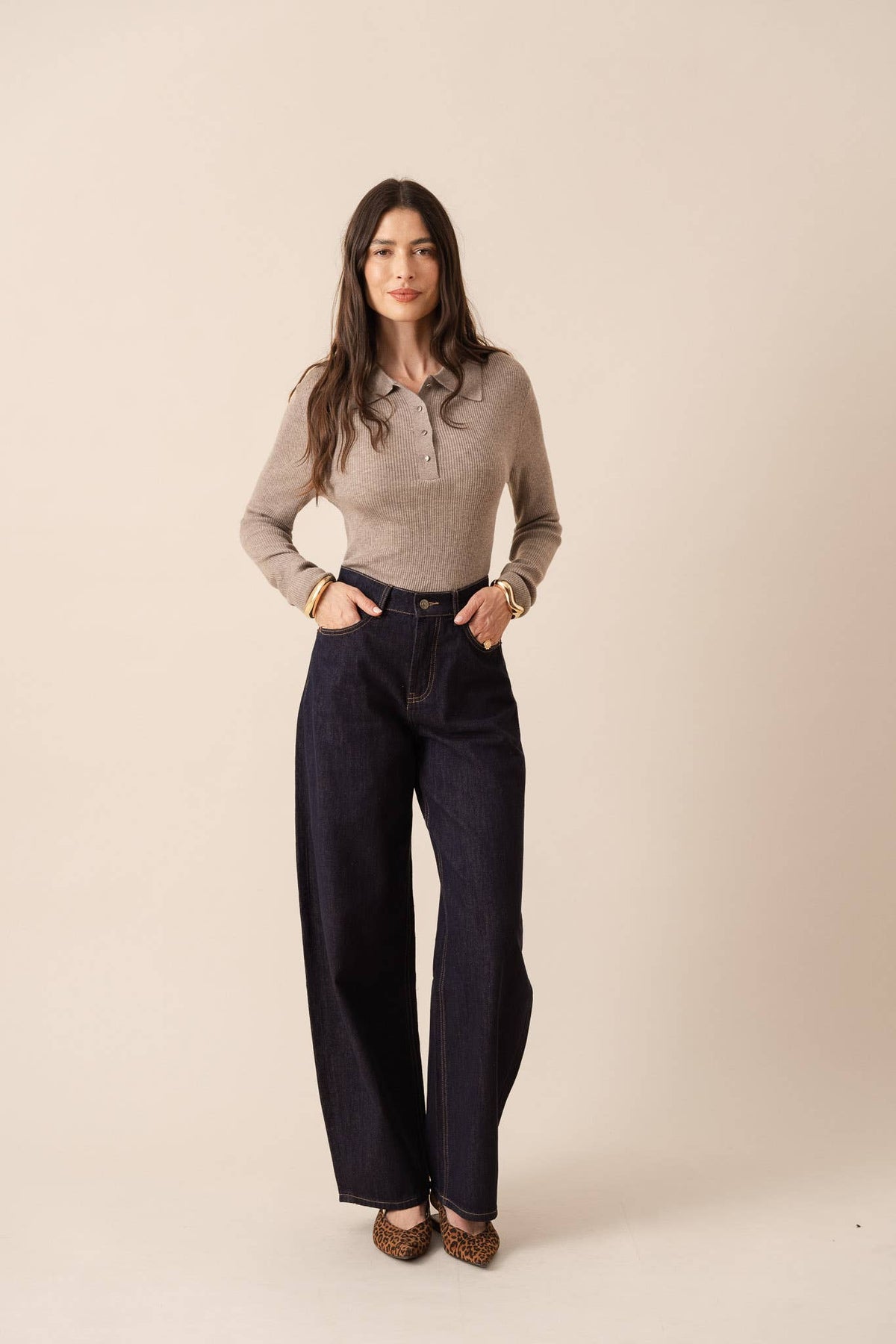 A woman stands against a plain beige studio background wearing a beige long-sleeve polo shirt, dark wide-leg denim jeans, and brown shoes.