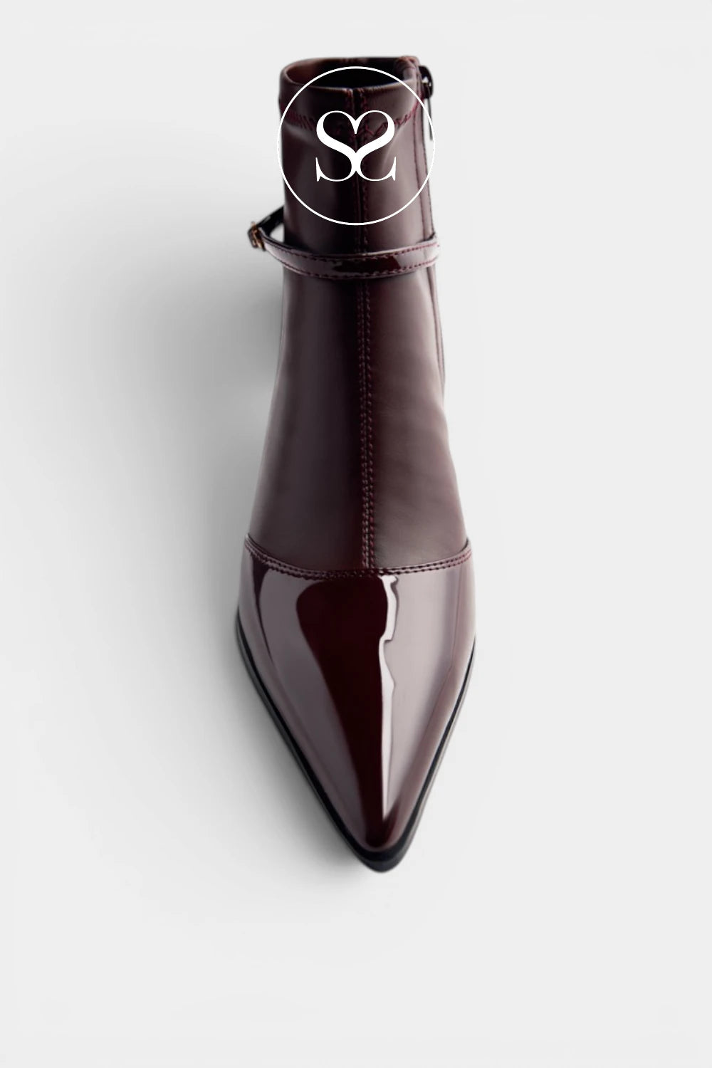 A dark brown patent leather ankle boot with a pointed toe and a side strap detail, shown from a top-down angle on a light gray background.