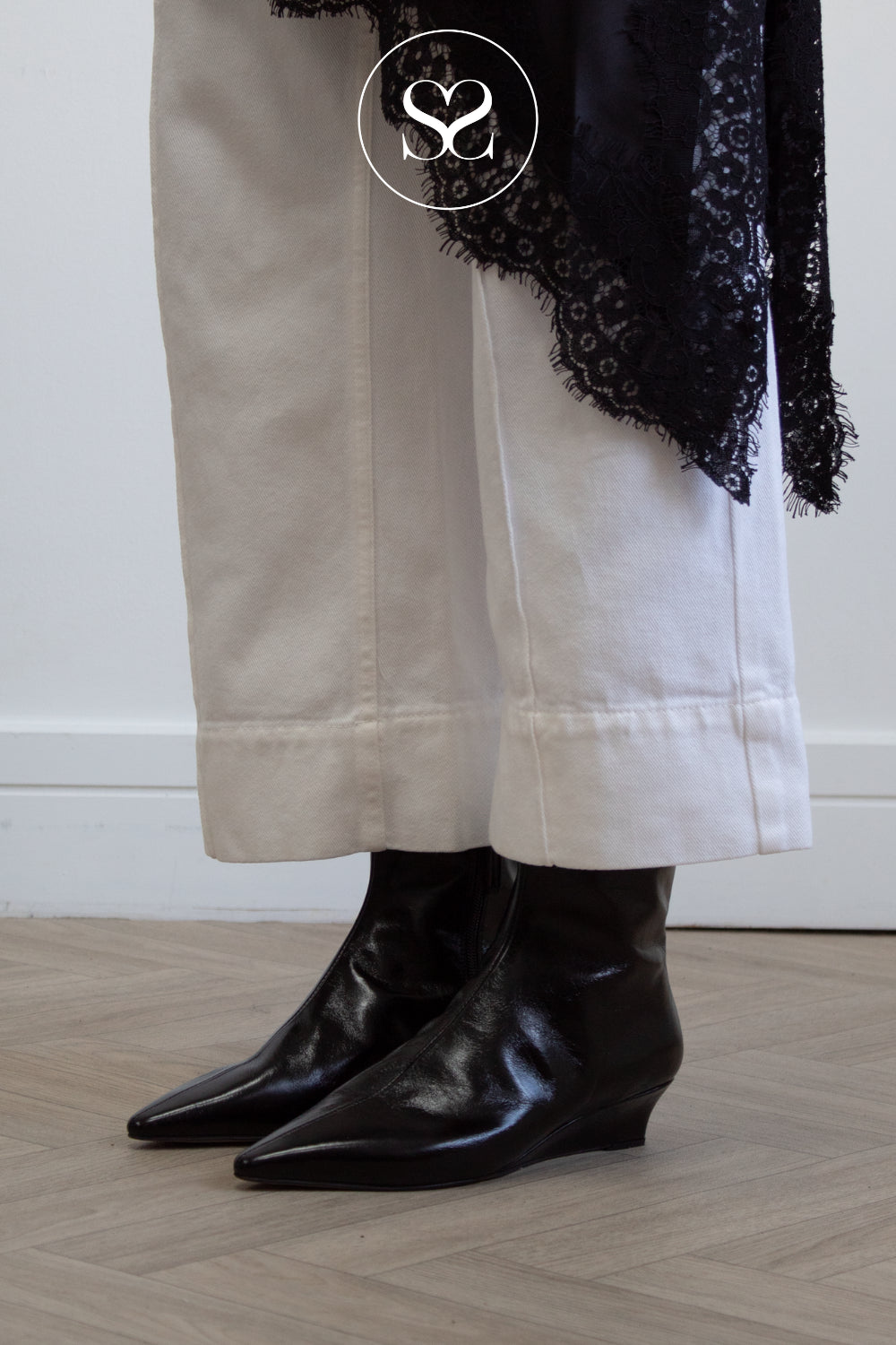 A close-up of a woman's legs wearing black high shine pointed-toe leather ankle boots with low wedge heels, paired with off-white cropped trousers and a black lace-trimmed garment draped over the hip, standing on a light wood floor.