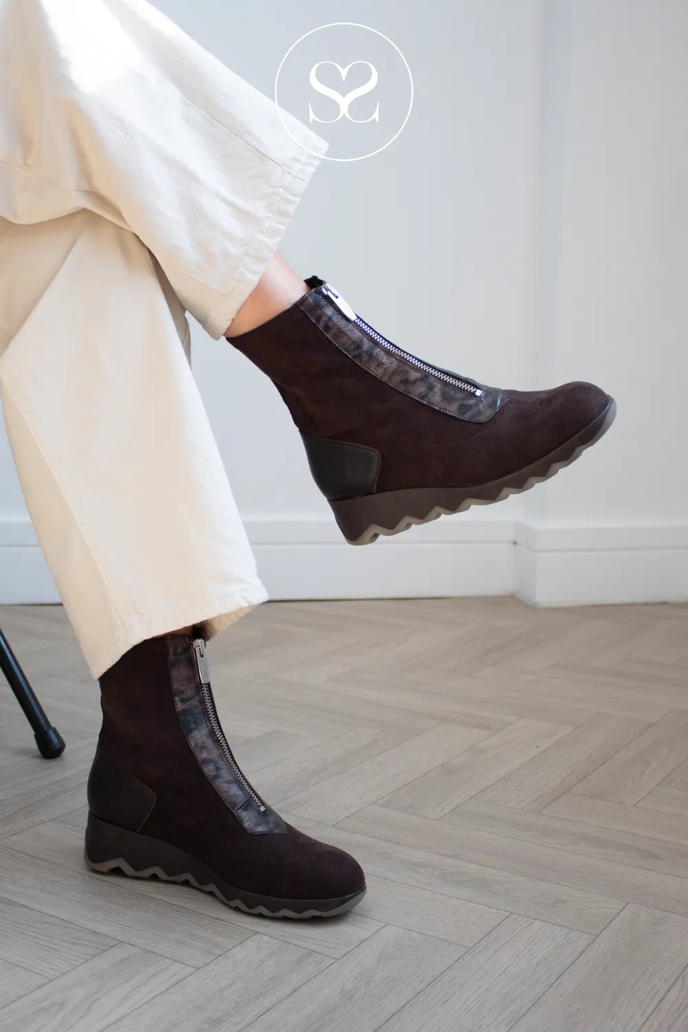 A person wearing Wonders dark brown wedge ankle boots with patterned elastic side panels and cream wide-leg trousers, standing on a light wood herringbone floor.