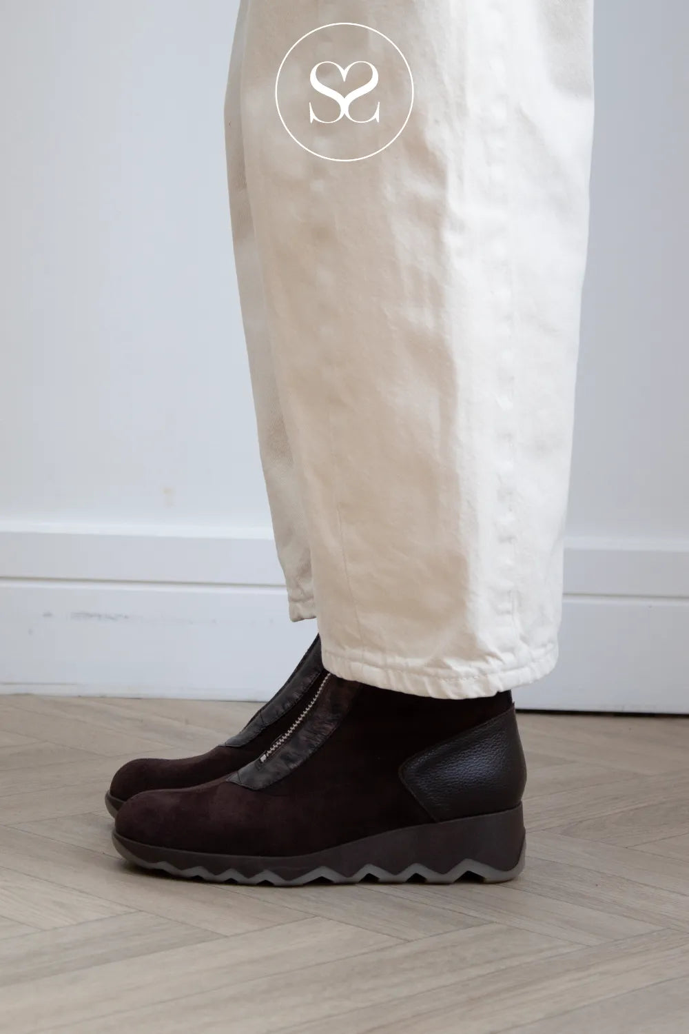 A person wearing Wonders dark brown Wedge ankle boots with a wavy sole and cream-colored trousers, standing on a light wood floor against a white wall.