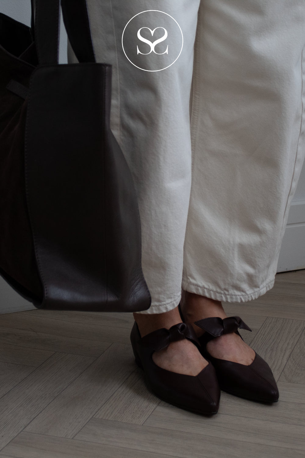 A close-up lifestyle shot of a person wearing dark brown pointed-toe flats with bow details, paired with off-white trousers and holding a large black leather tote bag. A circular 'SS' Sheneil Shoes logo is visible at the top.