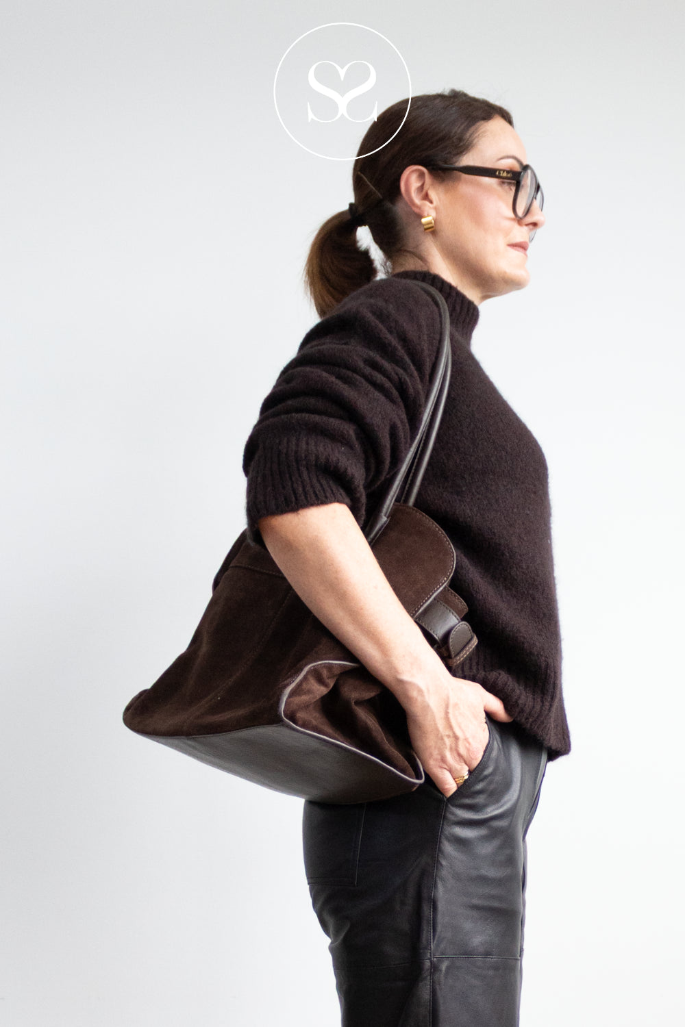 A woman in a dark brown knit sweater and black pants is shown in profile carrying a large dark brown leather Depeche shoulder bag against a light grey studio background in Ireland