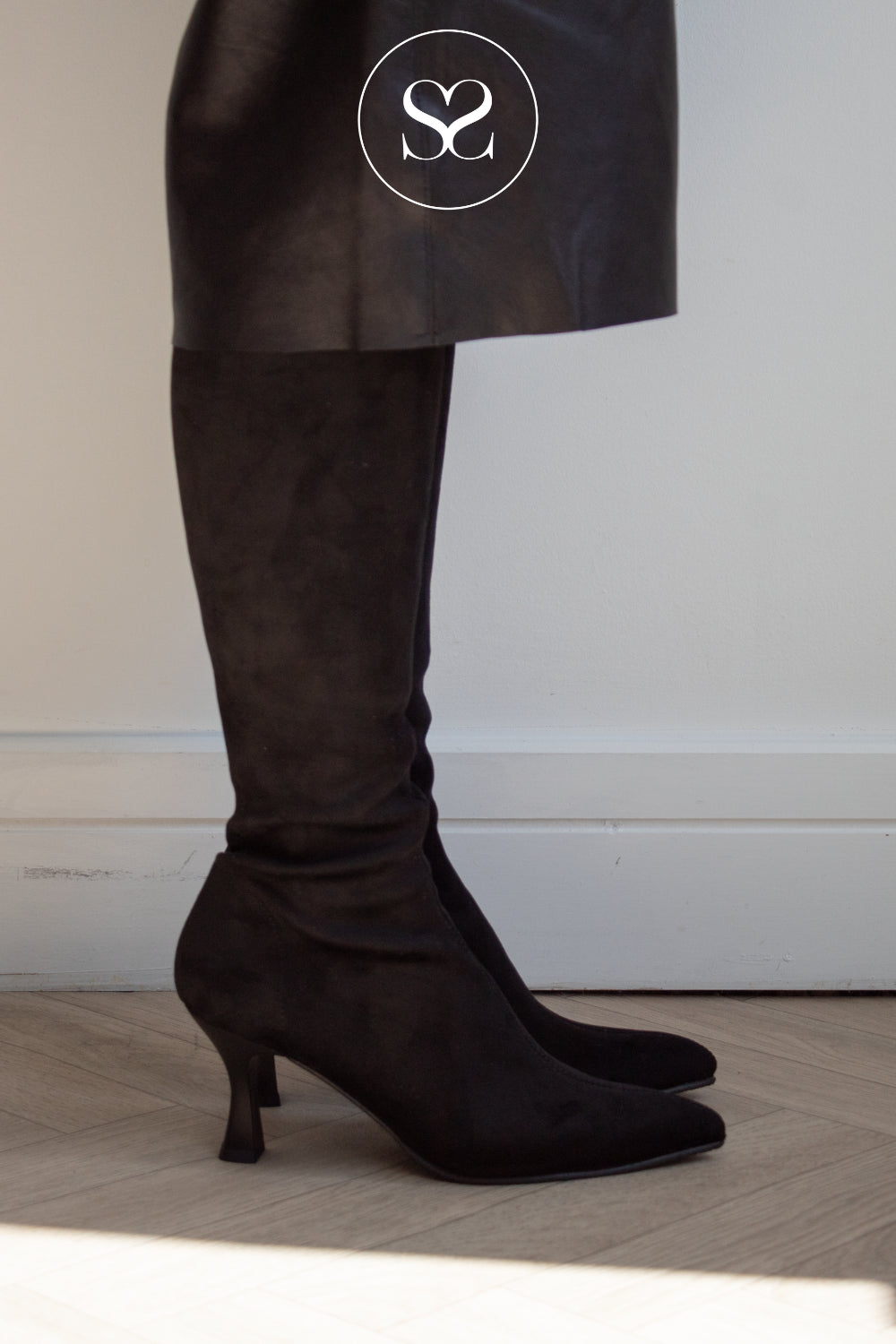 Black suede knee high sock boots with a pointed toe and kitten heel from Miss Elastic shown on a model wearing a black skirt. A white circular logo with 'SS' is visible on the skirt.