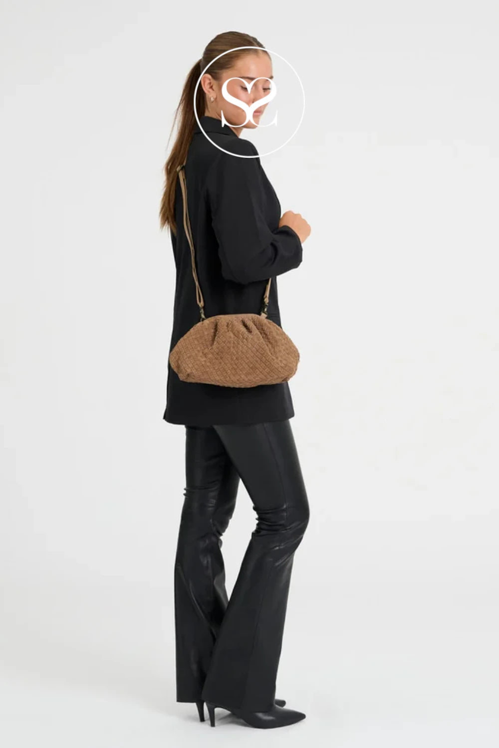 A woman in a black outfit is shown from the side against a light gray studio background, wearing a brown textured shoulder bag with a thin strap.