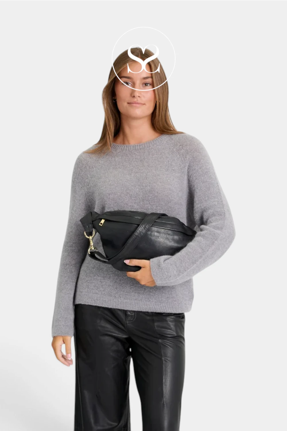 A woman modeling a black leather fanny pack with gold hardware, wearing a gray knit sweater and black leather pants against a light gray background.
