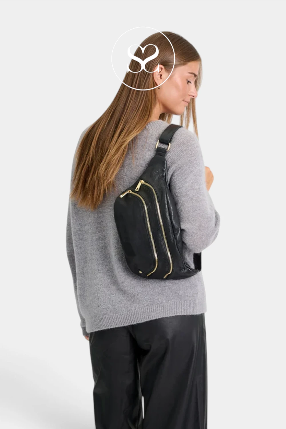 A woman wearing a gray knit sweater and black pants is shown from the back while wearing a black sling backpack with gold zipper accents. The background is a clean, light gray studio backdrop.