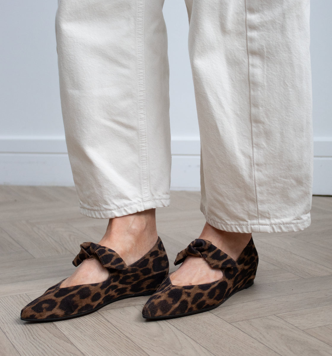 A woman wearing cream-colored wide-leg jeans and brown leopard-print ballet flats with bow details, standing on a light wood herringbone floor against a white wall taken in Galway, Ireland