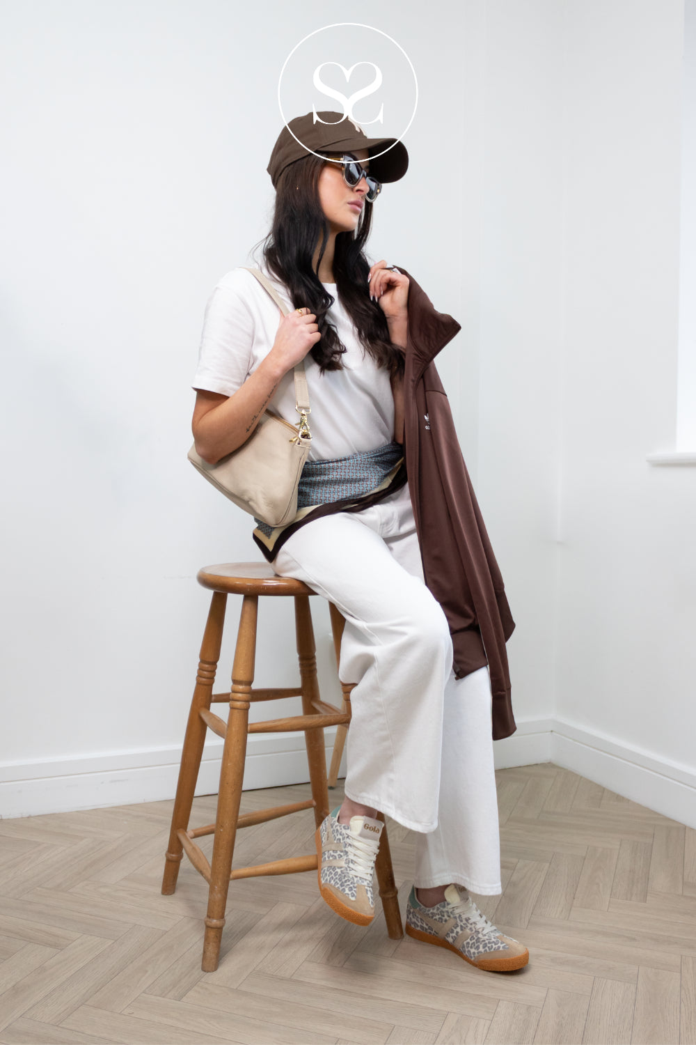 Woman sitting on a wooden stool wearing a white outfit with a brown jacket and gola leopard print sneakers.