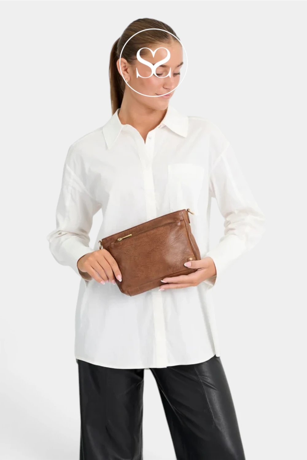 A woman wearing a white button-up shirt and black pants holds a brown faux-leather clutch bag with gold-tone hardware against a light gray studio background.