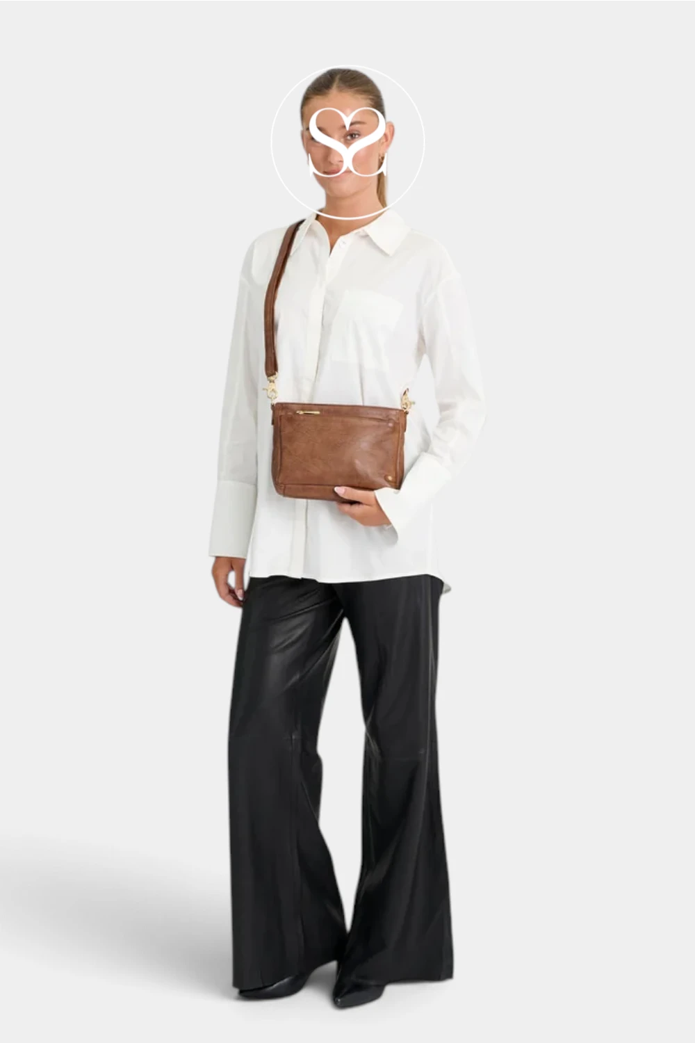 A model wearing a white button-down shirt, black wide-leg pants, and a brown leather crossbody bag with a gold logo, set against a plain light gray background.