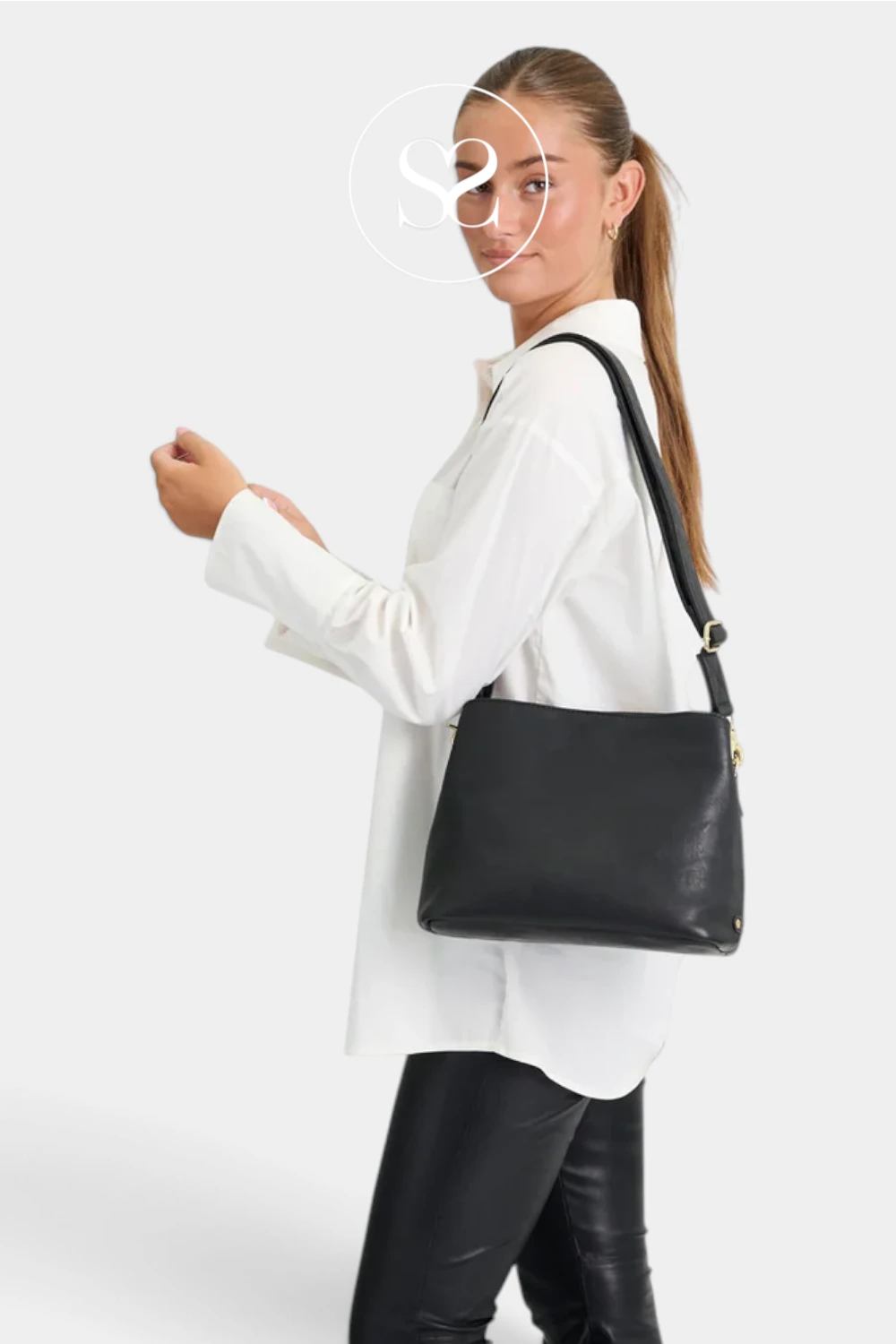 A woman wearing a white button-up shirt and black leather pants carries a black faux-leather shoulder bag with gold-tone hardware against a light gray studio background.