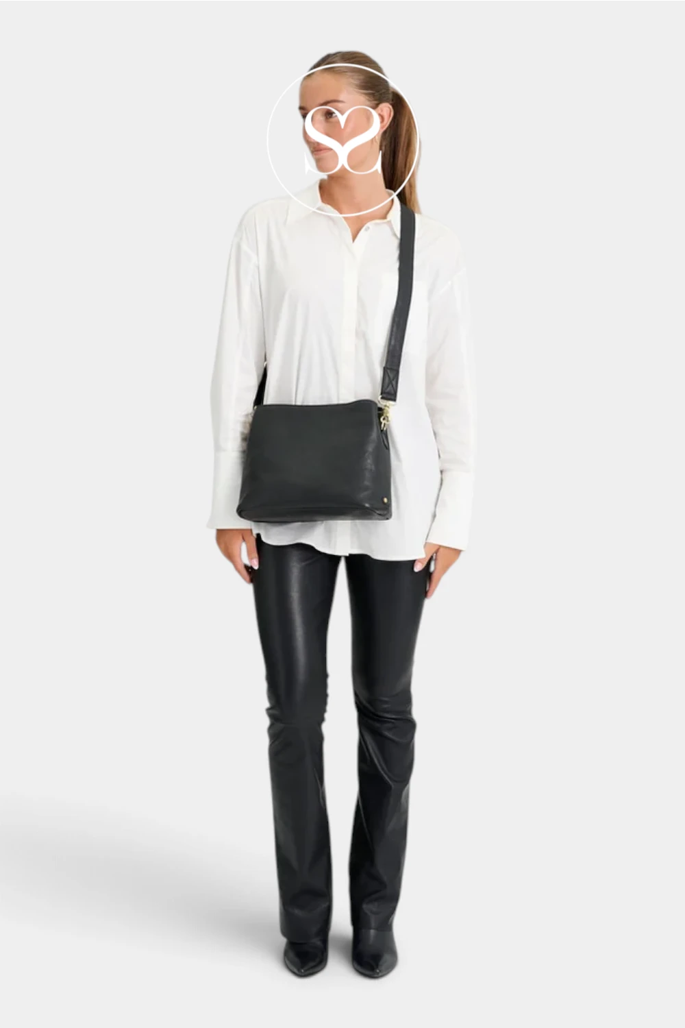 A woman modeling a black leather crossbody bag with gold hardware, worn over a white button-down shirt and black leather pants, against a light gray studio background.