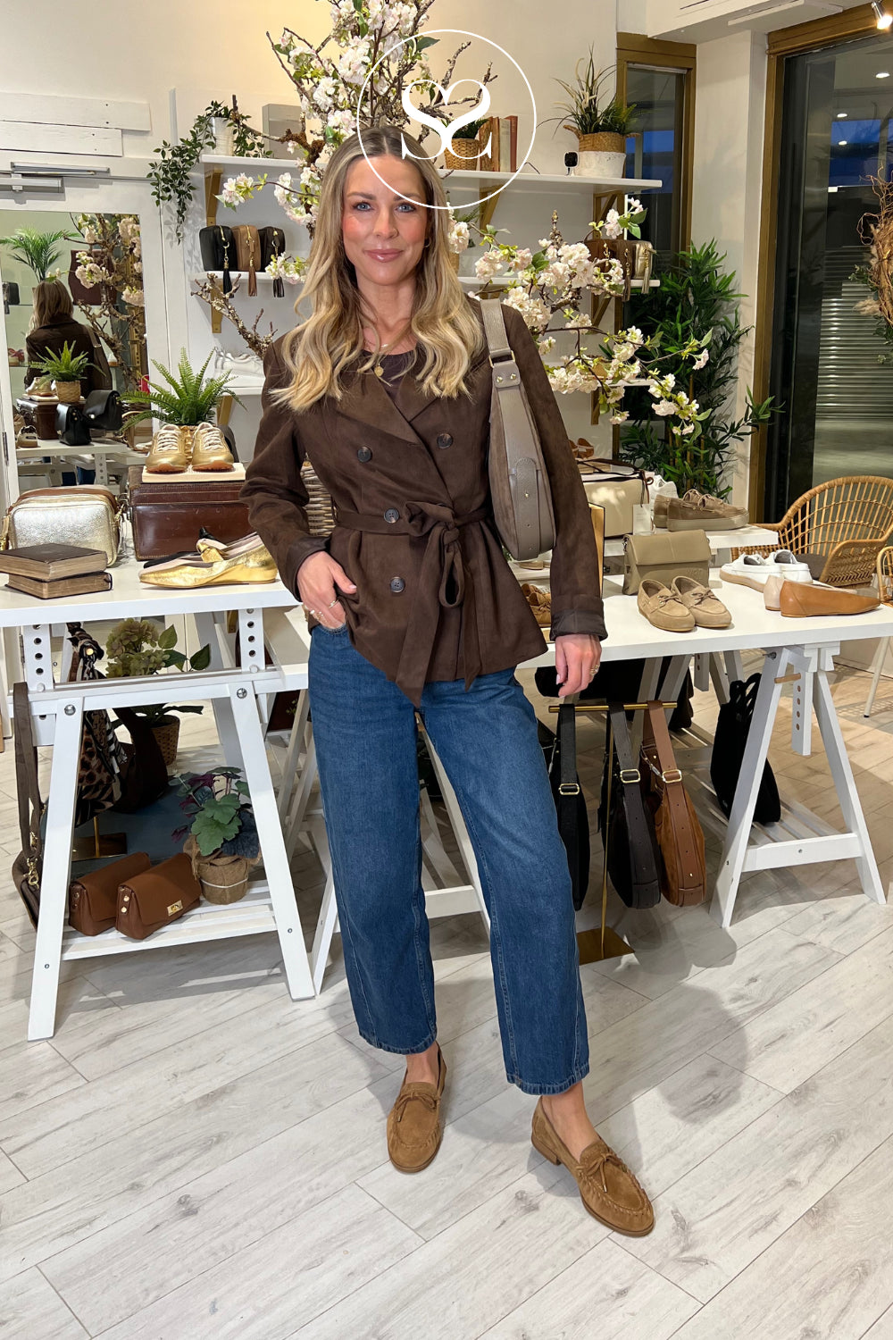 Woman in a brown trench coat and blue jeans standing in a store wearing unisa tan shoes