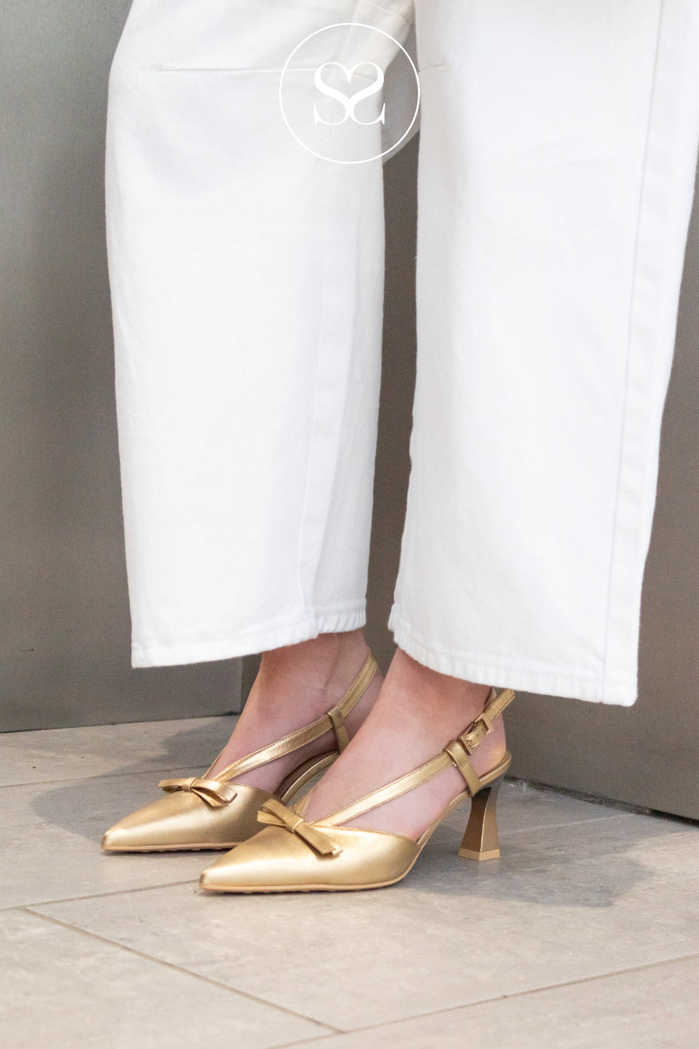 Gold high-heeled shoes from Hispanitas worn with white pants on a neutral background