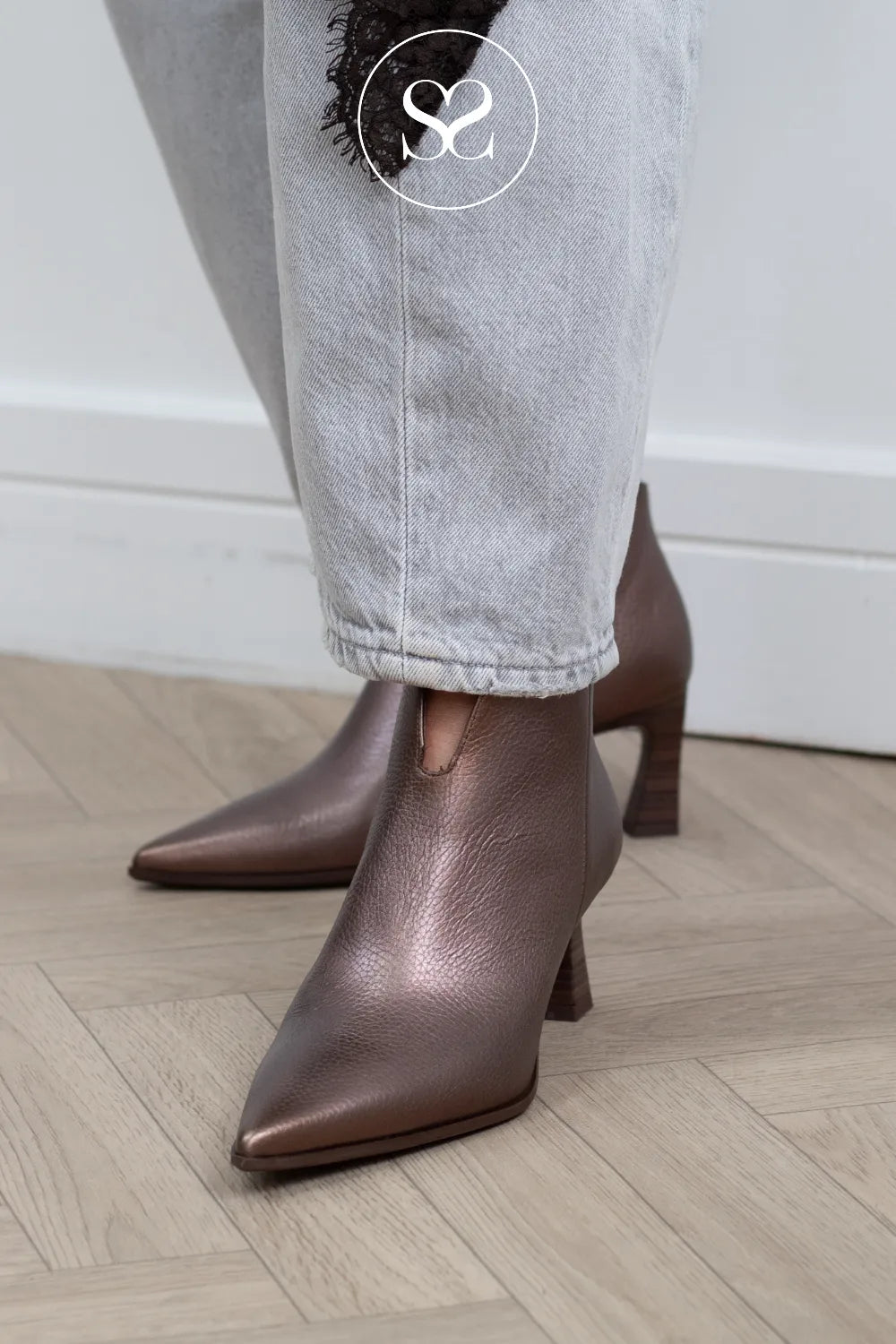 A person wearing pointed-toe metallic brown ankle boots with block heels, paired with light gray jeans, standing on a light wood herringbone floor.
