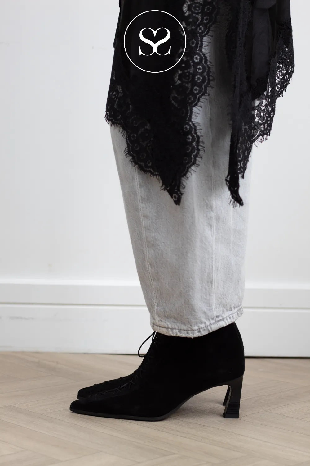 A person wearing black pointed-toe heeled ankle boots with fringe, paired with light gray jeans and a black lace shawl, standing on a light wood floor against a white wall.