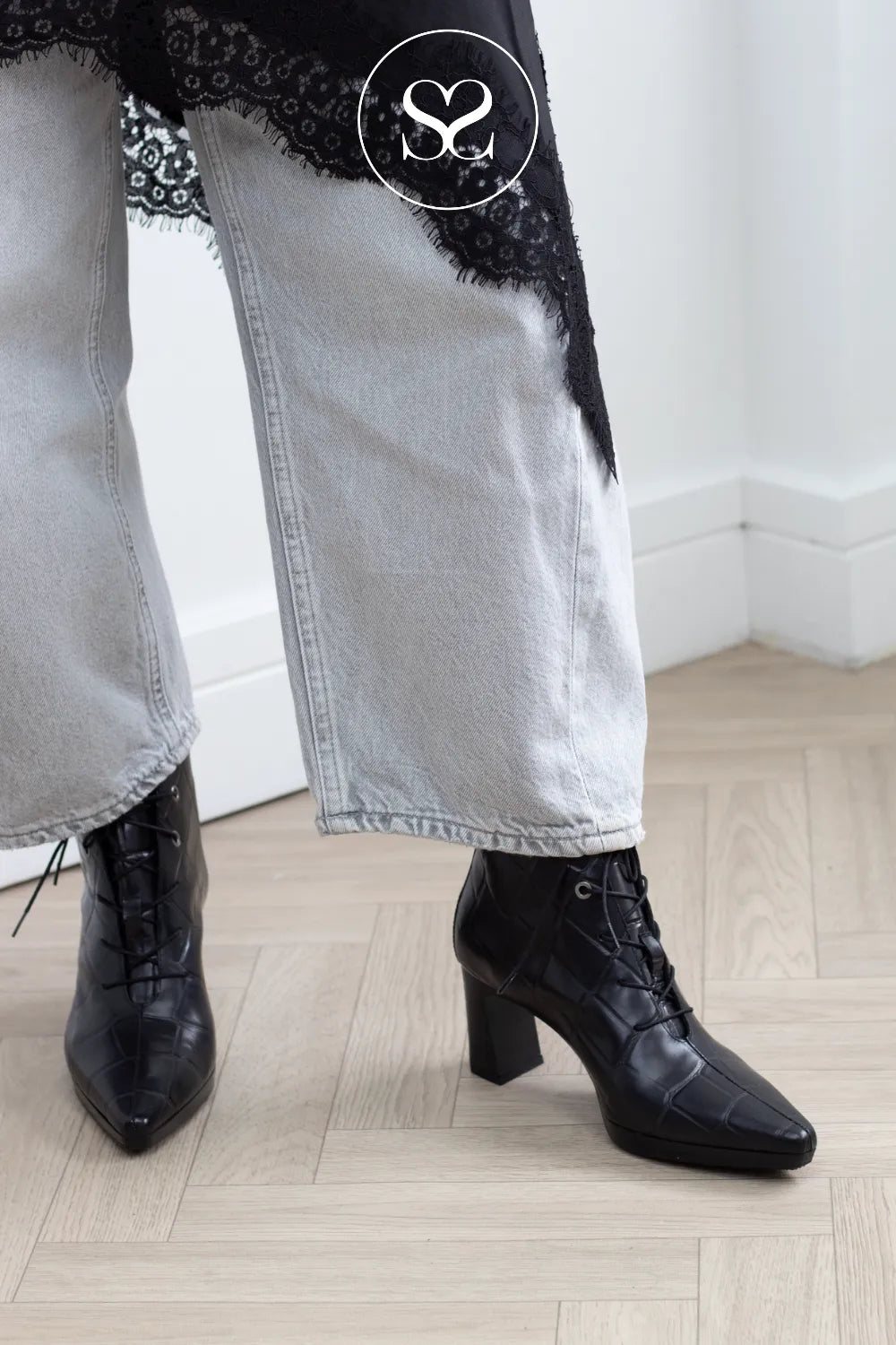 A close-up of a person's legs wearing black lace-up ankle boots with a block heel, paired with light-wash wide-leg jeans featuring black lace trim, standing on a light wood herringbone floor.