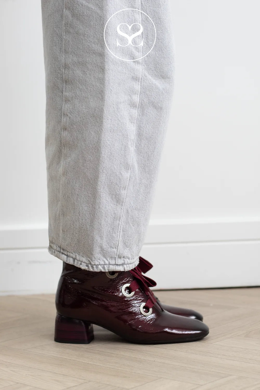 A person wearing light gray jeans and glossy dark burgundy ankle boots with a low block heel and decorative bow detail, standing on a light wood floor against a white wall.