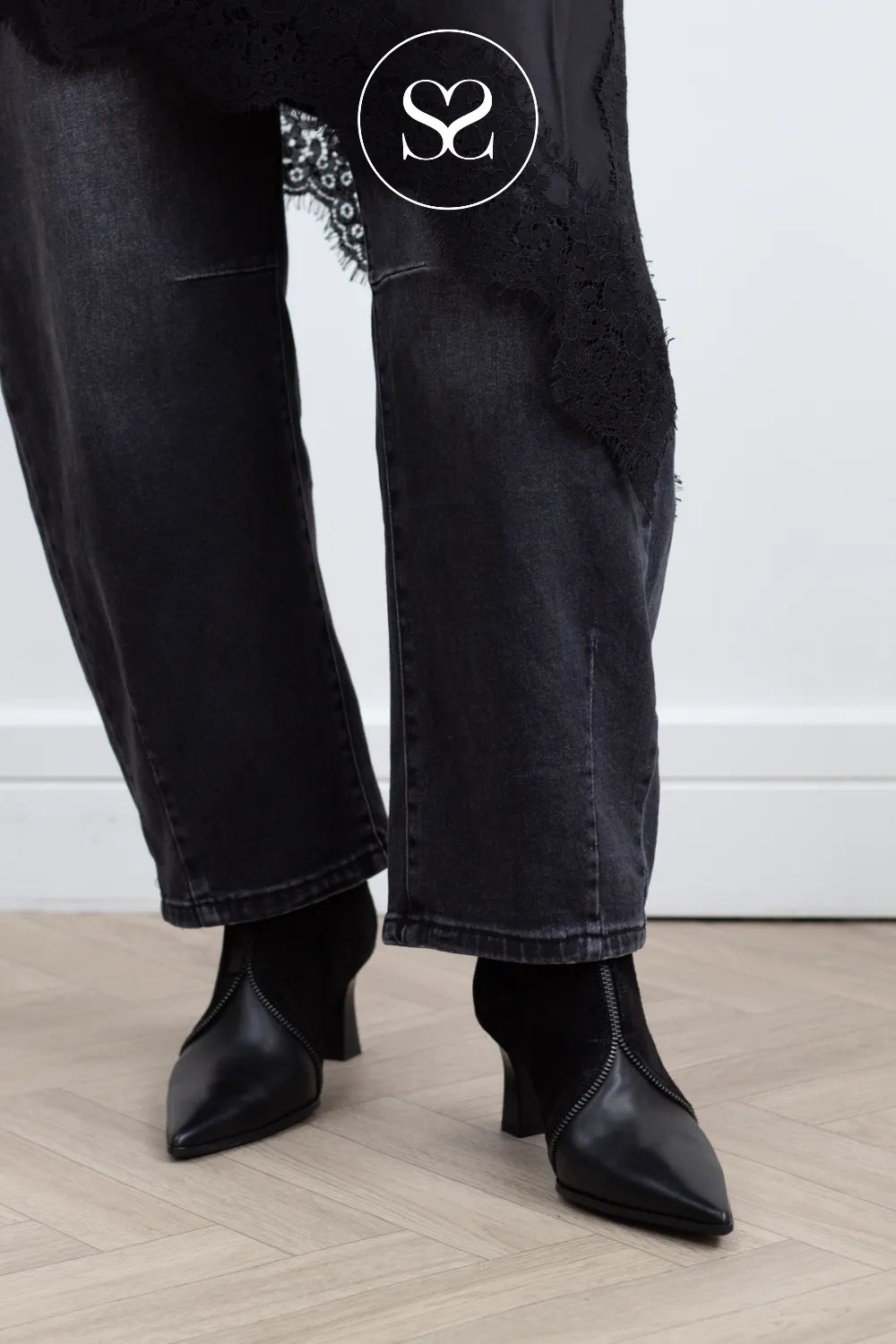 A person wearing wide-leg black jeans with lace and tassel embellishments, paired with black pointed-toe ankle boots, standing on a light wood floor against a white wall.
