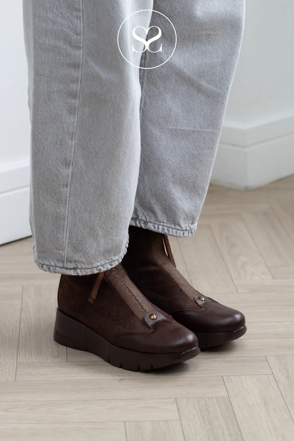 A person wearing dark brown leather slip-on platform ankle boots with light gray jeans, standing on a light wood herringbone floor.