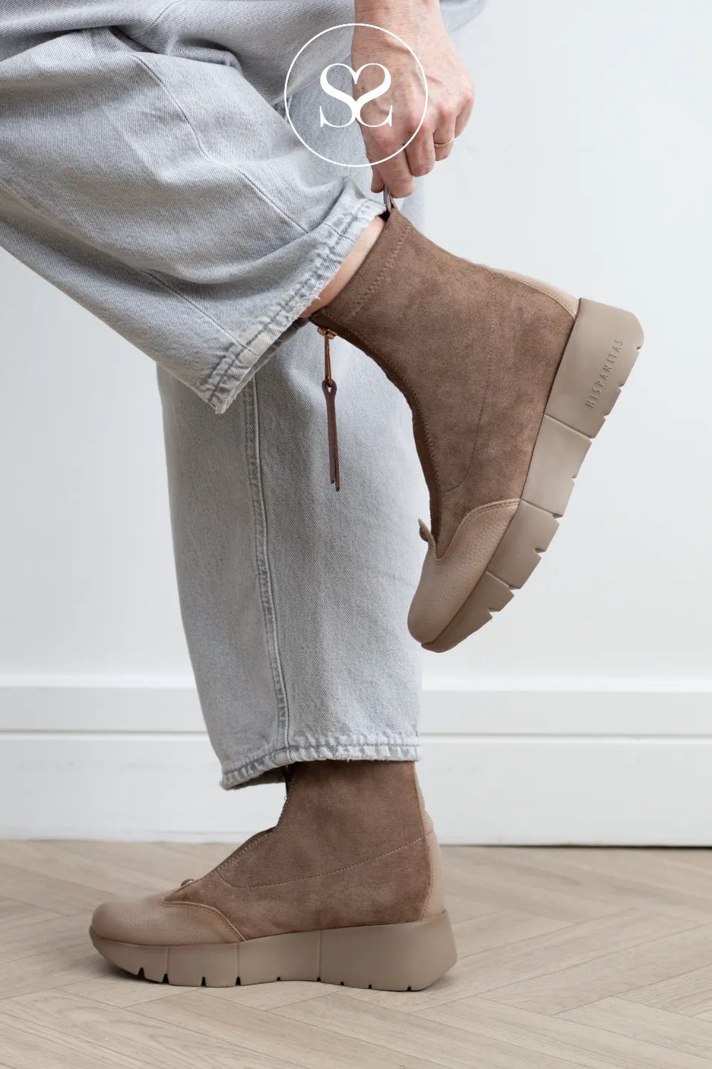 A person wearing light gray jeans and taupe-brown slip-on platform ankle boots, shown from the waist down against a clean white wall and light wood floor.