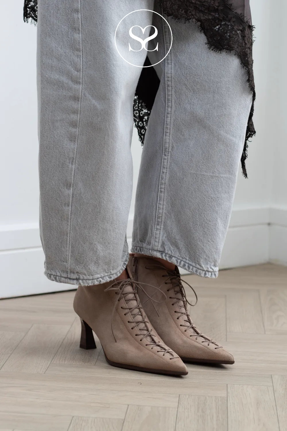A close-up of a person's legs wearing light gray wide-leg jeans, a sheer black lace top, and taupe pointed-toe lace-up ankle boots with a low block heel, standing on a light wood floor.