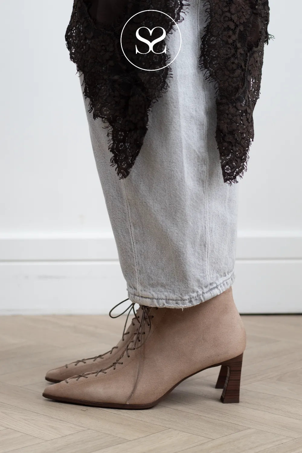 A person wearing pointed-toe beige lace-up ankle boots with a block heel, paired with light-wash jeans and a black lace draped garment, standing on a light wood floor against a white wall.