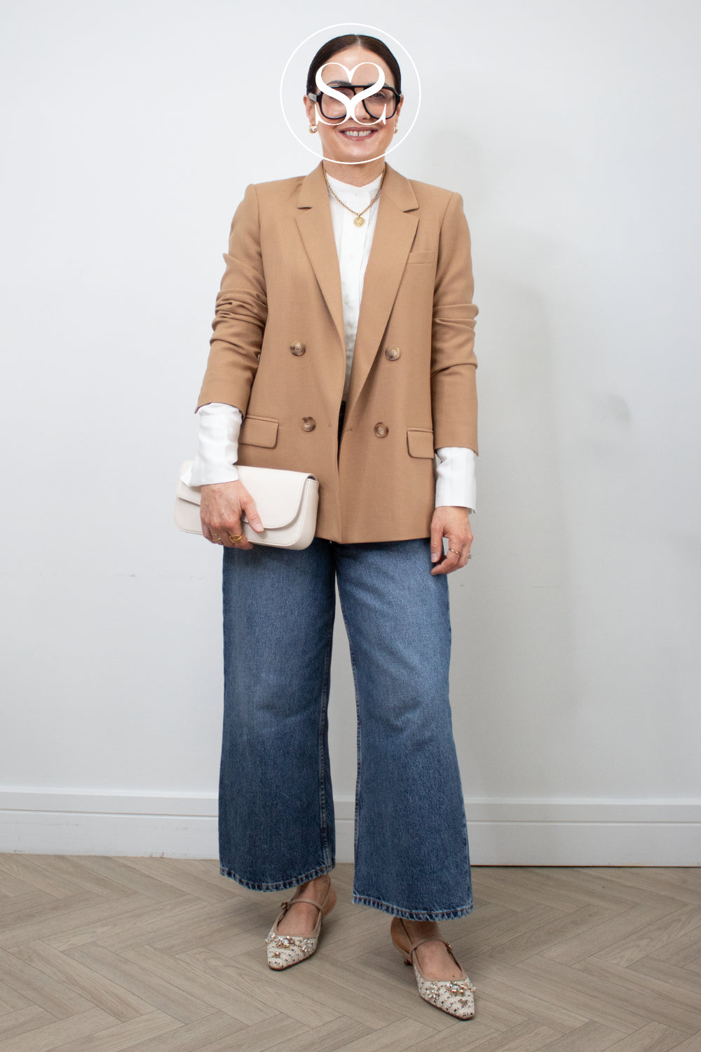 Woman wearing a beige blazer, white shirt, blue jeans, and embellished pointed toe kitten heels from lodi with an Ivory clutch bag on a plain background