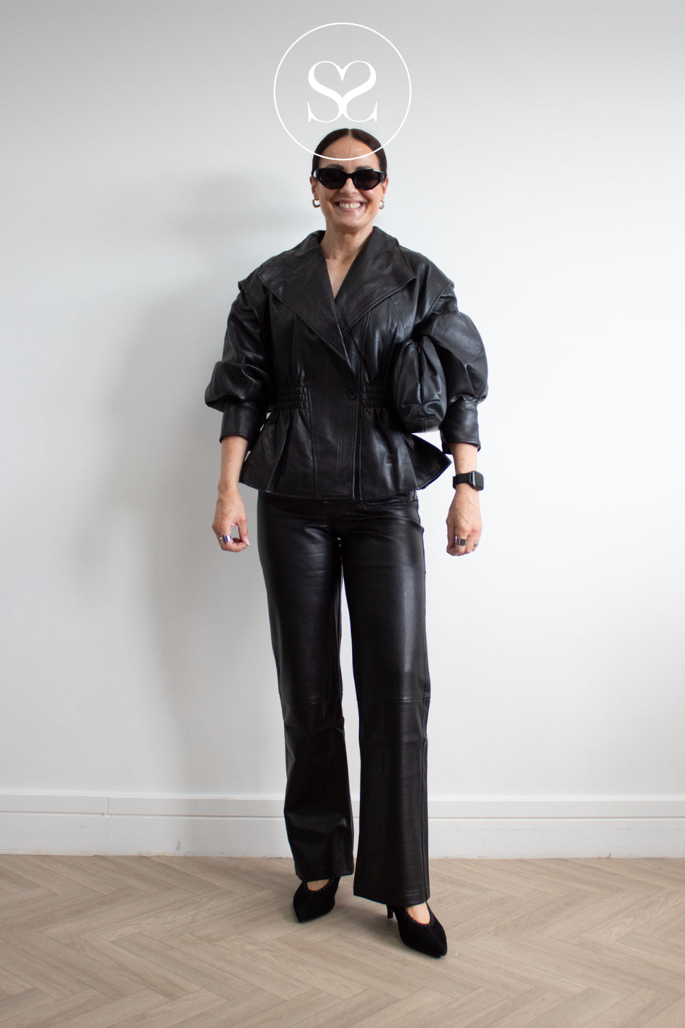 A woman modeling a black faux leather jacket and matching wide-leg trousers, styled with black sunglasses, a black watch, and black pointed lodi kitten heel shoes, standing against a white wall with a light wood herringbone floor.