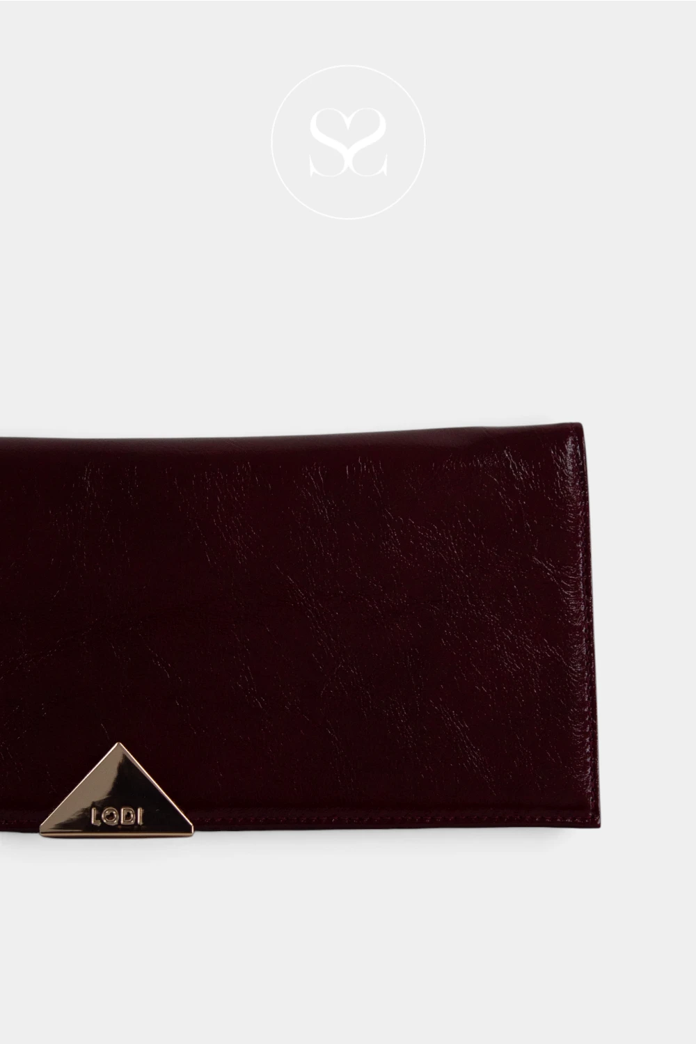 A close-up of a dark burgundy leather evening clutch bag with a gold-tone triangular metal accent on the front, set against a light grey background with a faint sheneil shoes logo watermark at the top. Bag from Lodi Available in Ireland