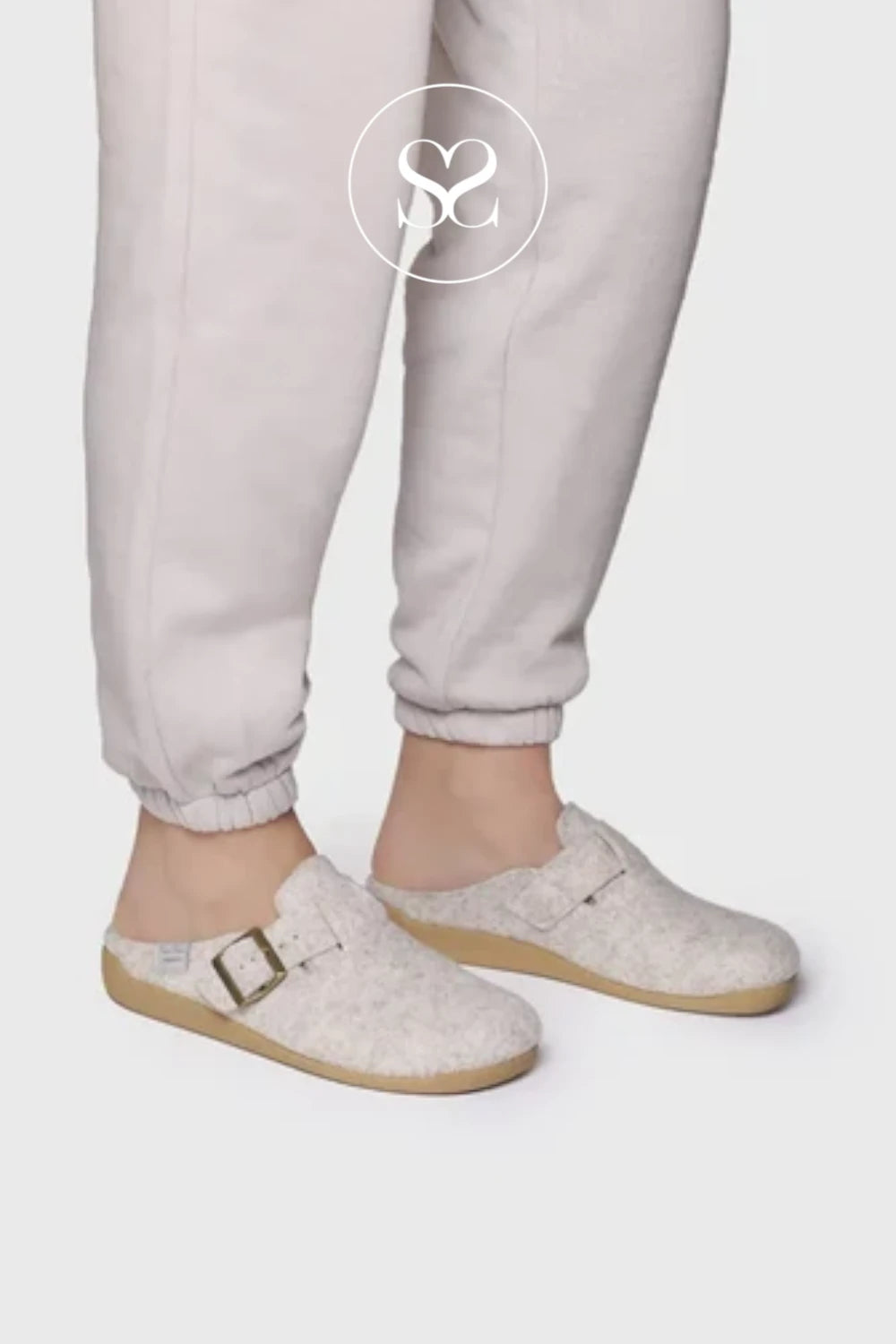 A person wearing light beige jogger pants and matching textured clog-style slip-on shoes with a small buckle detail, shown against a plain light gray studio background. A white circular logo watermark is visible near the upper leg.