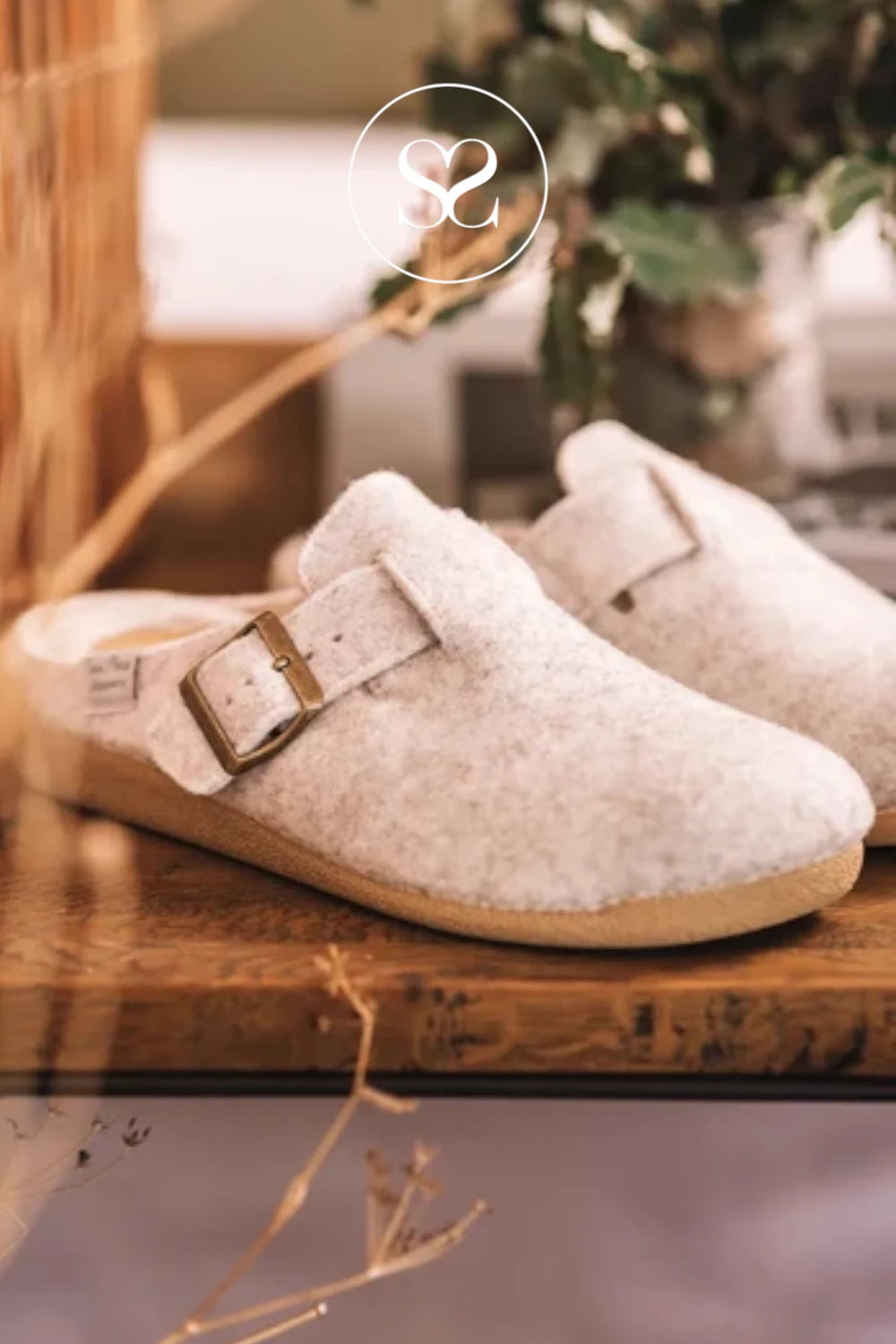 A pair of off-white felt clogs with adjustable brown straps and gold-tone buckles, resting on a rustic wooden surface with a warm, softly blurred background.